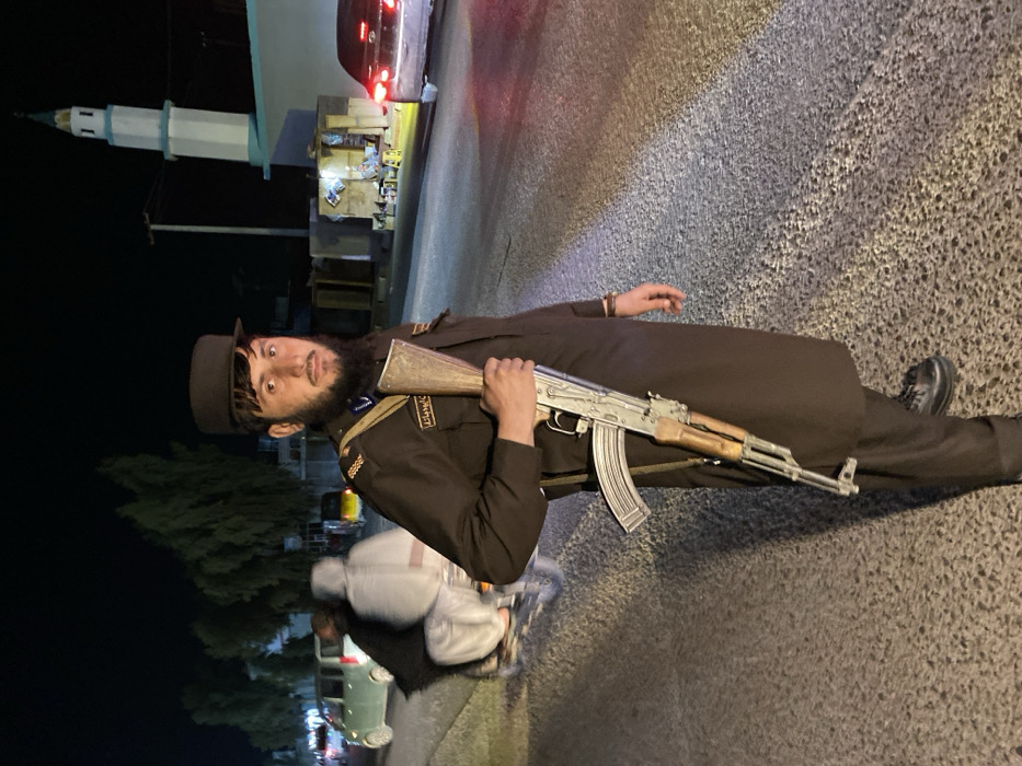 A nighttime street scene in Kandahar, Afghanistan, captures a young adult male in a dark brown uniform, black cap, and black shoes, standing on a paved road and looking directly at the camera. He wears gold-colored epaulets and a badge with Arabic script on his left chest, along with a dark blue shoulder strap. He possesses a dark beard and mustache. The man is holding an AK-pattern rifle, featuring a wooden foregrip and stock with a ribbed steel magazine, vertically with the buttstock resting near the ground, supported by his right hand.
In the left background, a light-colored van is visible behind a motorcycle carrying two blurred individuals, one in dark clothing and one in light clothing, both moving from left to right. In the right background, a dark-colored sedan with illuminated taillights is parked or moving slowly. Adjacent to the sedan is a small, brightly lit street stall with items displayed and two small stools in front. Further right and behind the stall stands a white mosque featuring a tall, slender minaret topped with a light blue-green conical spire and balconies. Dark trees are visible against the night sky in the distant left background. The scene is illuminated by artificial light sources from vehicles, the street stall, and ambient streetlights.