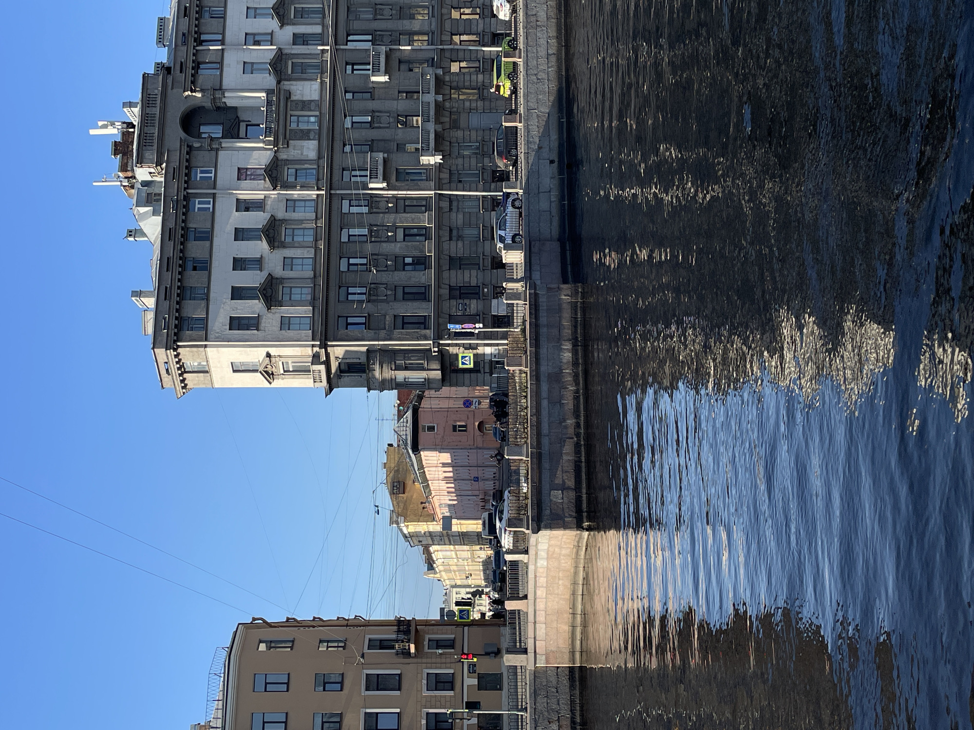 The image displays an urban scene in Saint Petersburg, Russia, specifically at the intersection of the Griboyedov Canal and Gorokhovaya Street, viewed looking southeast. The foreground is dominated by the dark blue, rippled water of the Griboyedov Canal, which reflects parts of the surrounding architecture and the clear blue sky.
On the right bank of the canal stands a large, multi-story building, primarily grey and white, featuring an ornate classical facade with numerous windows, balconies, pilasters, and a central arched section topped with a small figure. The rooftop displays various structures, including several white cellular antennas. A road runs along this embankment, separated from the canal by a stone wall and a black metal railing. Parked or moving vehicles on this road include a silver SUV, a dark sedan, and a lime-green compact car.
On the left, a multi-story, beige corner building with dark-framed windows and balconies is visible. A red traffic light is positioned near this building. Further down the street, identified as Gorokhovaya Street, various buildings line the thoroughfare, including a distinctive multi-story pink building in the mid-distance, flanked by yellow and lighter-toned structures. Overhead wires crisscross the expanse of the clear blue sky above the street. Small groups of pedestrians are visible on both canal embankments and along Gorokhovaya Street. The scene is illuminated by strong, direct sunlight, creating distinct shadows on the building facades.