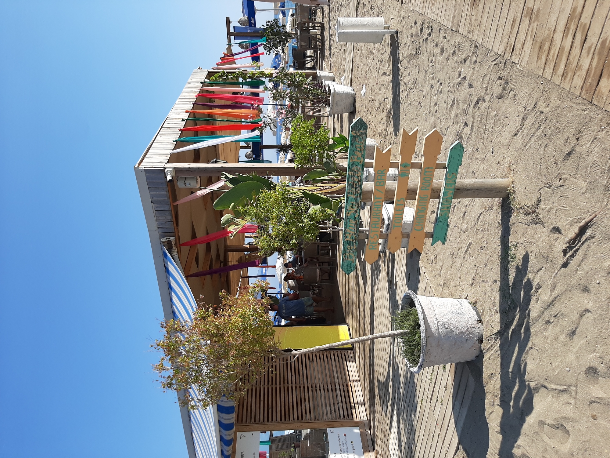 The image depicts a beachside establishment in Alanya, Turkey, featuring a wooden structure with a striped awning providing shade. Colorful fabric streamers hang from the roof. A small tree in a white pot stands on a wooden walkway. In the foreground, a wooden signpost with multiple arrow-shaped signs directs to "RESTAURANT/BAR", "TOILETS", "CHANGING ROOM", and "SHOWERS". The ground is sandy, with footprints visible. In the background, tables and chairs are set up, and people are seated, suggesting a dining area. Beach umbrellas and the sea are visible further in the distance under a clear blue sky.