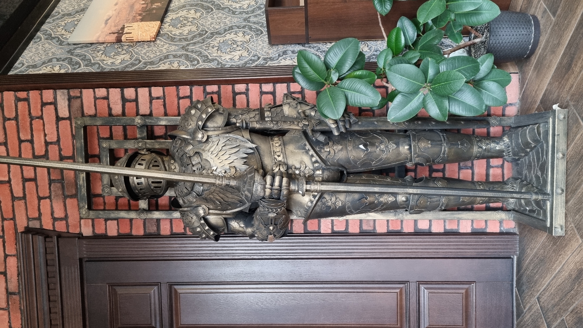 A life-sized suit of armor, resembling a medieval knight, stands upright in a room. The armor is made of a dark, metallic material with intricate details and is holding a long sword. Behind the armor, a brick wall forms the background. To the left of the armor is a dark wooden door with vertical paneling. To the right, a large potted plant with glossy green leaves is visible. In the background on the right, a framed picture depicting a castle is hung on a patterned wallpapered wall, and a dark wooden shelf unit is present. The floor is covered with wooden planks. The scene appears to be an indoor decoration, potentially in a commercial establishment or a residence, located in the city of Smila, Ukraine.