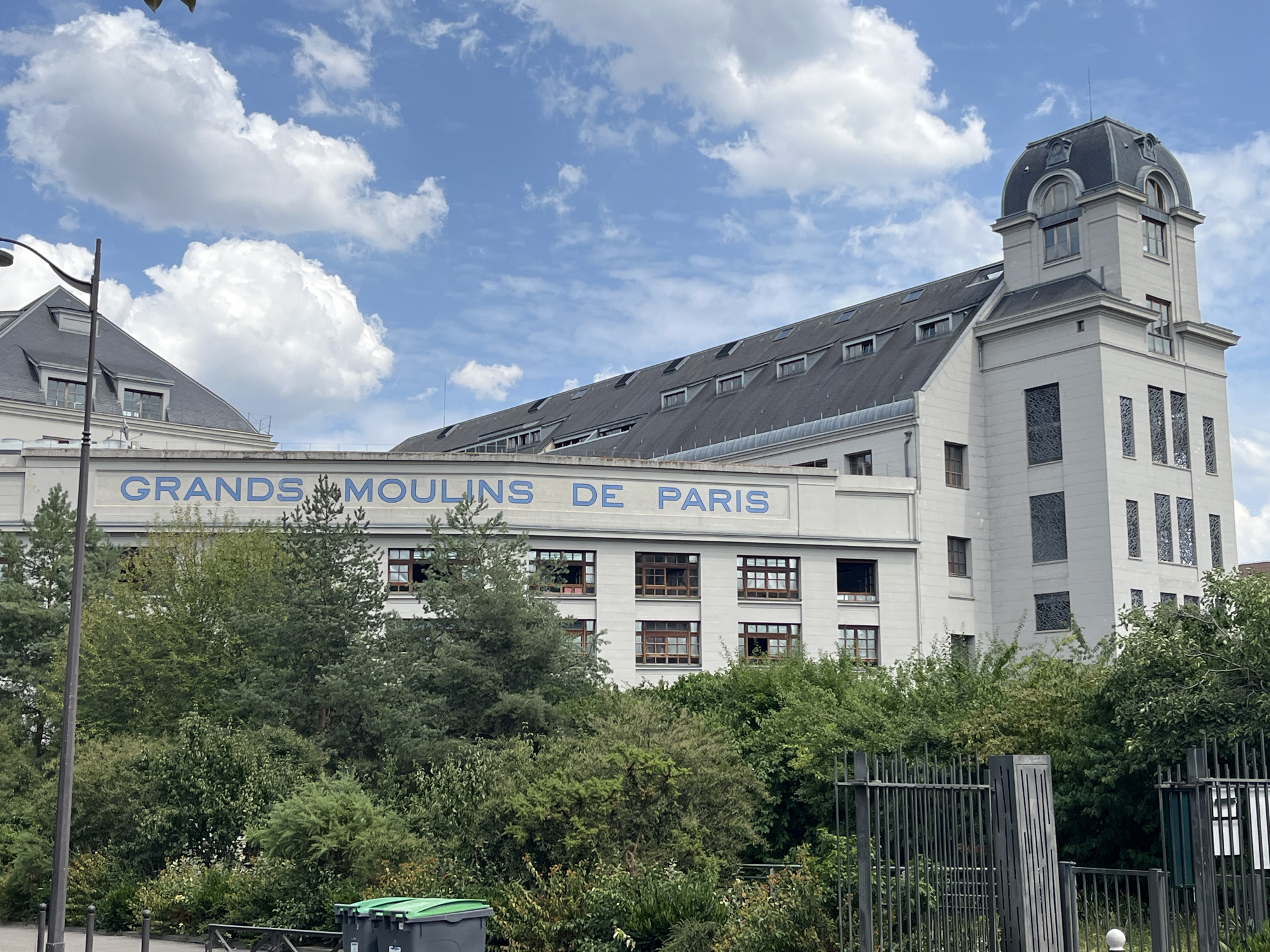 A slightly low-angle, medium shot captures a building with the words "GRANDS MOULINS DE PARIS" in blue lettering across its facade. The building is white with a gray, mansard-style roof dotted with dormer windows. A prominent tower with a domed roof is situated on the right side of the building. The building is surrounded by lush green trees and foliage. A black metal fence runs across the bottom of the image, with a green trash bin visible to the left. The sky is blue with scattered white clouds. The scene appears to be an exterior view of an industrial or historical building in an urban setting.