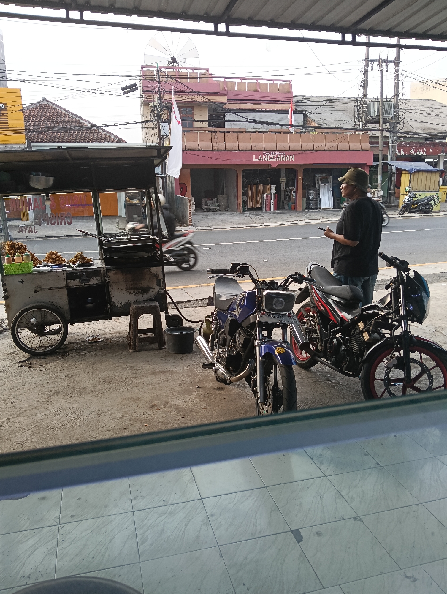 A man in a green cap and black shirt stands on a sidewalk, holding a phone, looking to his right. Two motorcycles are parked on the sidewalk near him. To the left, a food cart with a glass display case is filled with fried chicken. In the background, a building with red trim has signage that reads "TOKO KACA LANGEANAN". A white flag with red markings flies from a pole in front of the building. The scene appears to be in Desa Cipakat, Indonesia, as indicated by the context of the request. The sky is overcast, and electrical wires are visible overhead.
