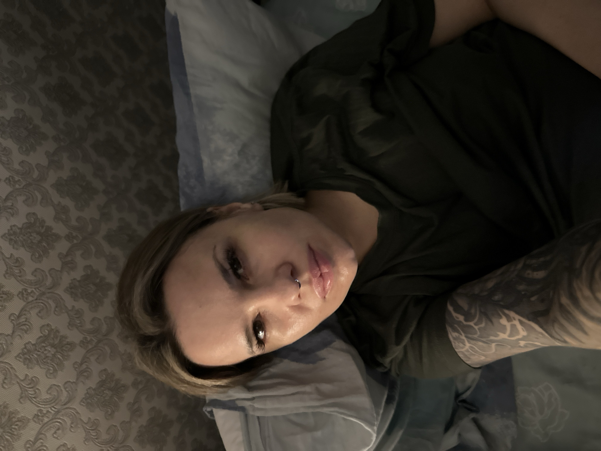 An indoor scene depicts a female subject lying on a bed or similar soft surface, observed from a high angle. The subject has light skin, medium-length blonde or light brown hair, and dark eyes with prominent eyelashes. A silver hoop septum piercing is visible on her nose. She wears a dark olive green short-sleeved t-shirt. Her right arm (viewer's left) displays a full sleeve tattoo composed of intricate black and grey patterns. Her head is positioned on a pillow featuring a blue and white striped pillowcase. The background shows a wall covered in light-colored wallpaper with a repetitive, ornate damask-style pattern. The lighting is dim, suggesting an interior environment. The precise location within Rostov-on-Don, Russia, is not identifiable from the visual content provided.