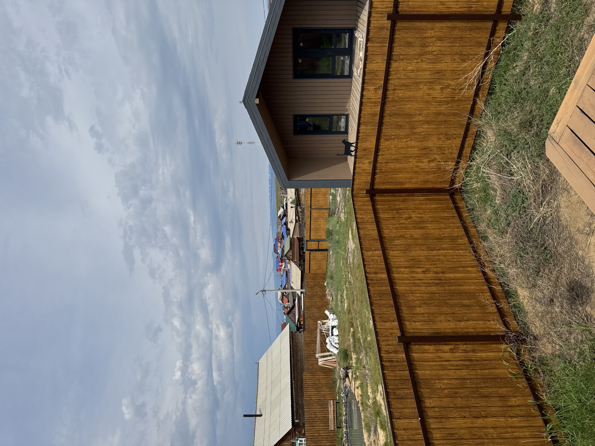 This is a landscape photograph taken in Khuzhir, Russia, depicting a residential area with a focus on fences and buildings under a cloudy sky. In the foreground, a tall, wooden fence with a dark metal frame runs diagonally across the frame, creating a sense of depth. Beyond this fence, another wooden fence encloses a yard with dry grass and some green patches.
To the left, a single-story wooden house with a white, corrugated metal roof and a visible chimney is situated. Adjacent to it is a wooden bench placed near a small body of water, possibly a pond. Several white sacks and wooden beams are also present in this area, suggesting ongoing construction or material storage.
In the middle ground, a cluster of houses with colorful roofs (red, blue, and green) are visible, indicating a village or small town setting. In the right portion of the frame, a modern, two-story house with a dark brown exterior and a dark gray roof stands out. This house features large windows and a wooden deck. A small black cat is perched on the deck railing of this modern house, facing left. Further in the background, a tall, slender tower, possibly a communication tower, is visible against the sky. The horizon shows rolling hills and a body of water, likely Lake Baikal, given the location. The sky is predominantly covered with light gray clouds, with some patches of blue peeking through.