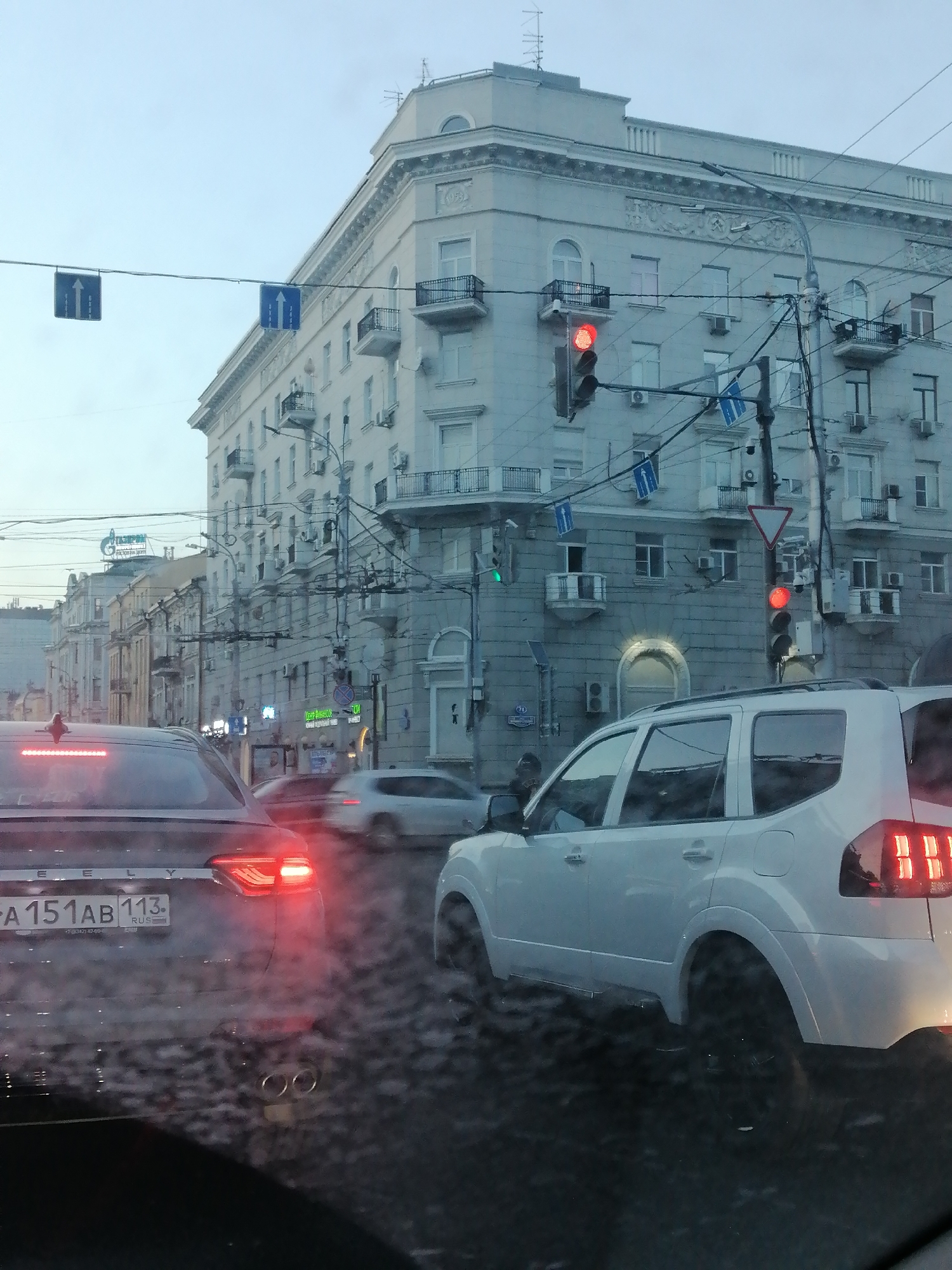 The image shows a street scene in Rostov-on-Don, Russia, likely during overcast weather. In the foreground, the rear of a dark-colored sedan is visible, with its red taillights illuminated. To the right of the sedan, a white SUV is also visible, also with its rear taillights illuminated. Both vehicles are stopped, presumably at a traffic light.
The background features a large, multi-story building with classical architectural elements, including balconies and decorative moldings. Several traffic signals and directional signs are suspended over the street. The overall impression is of an urban street with moderate traffic and typical city infrastructure. The presence of Russian license plates on the vehicles and the building's architecture suggest the location is indeed Russia.