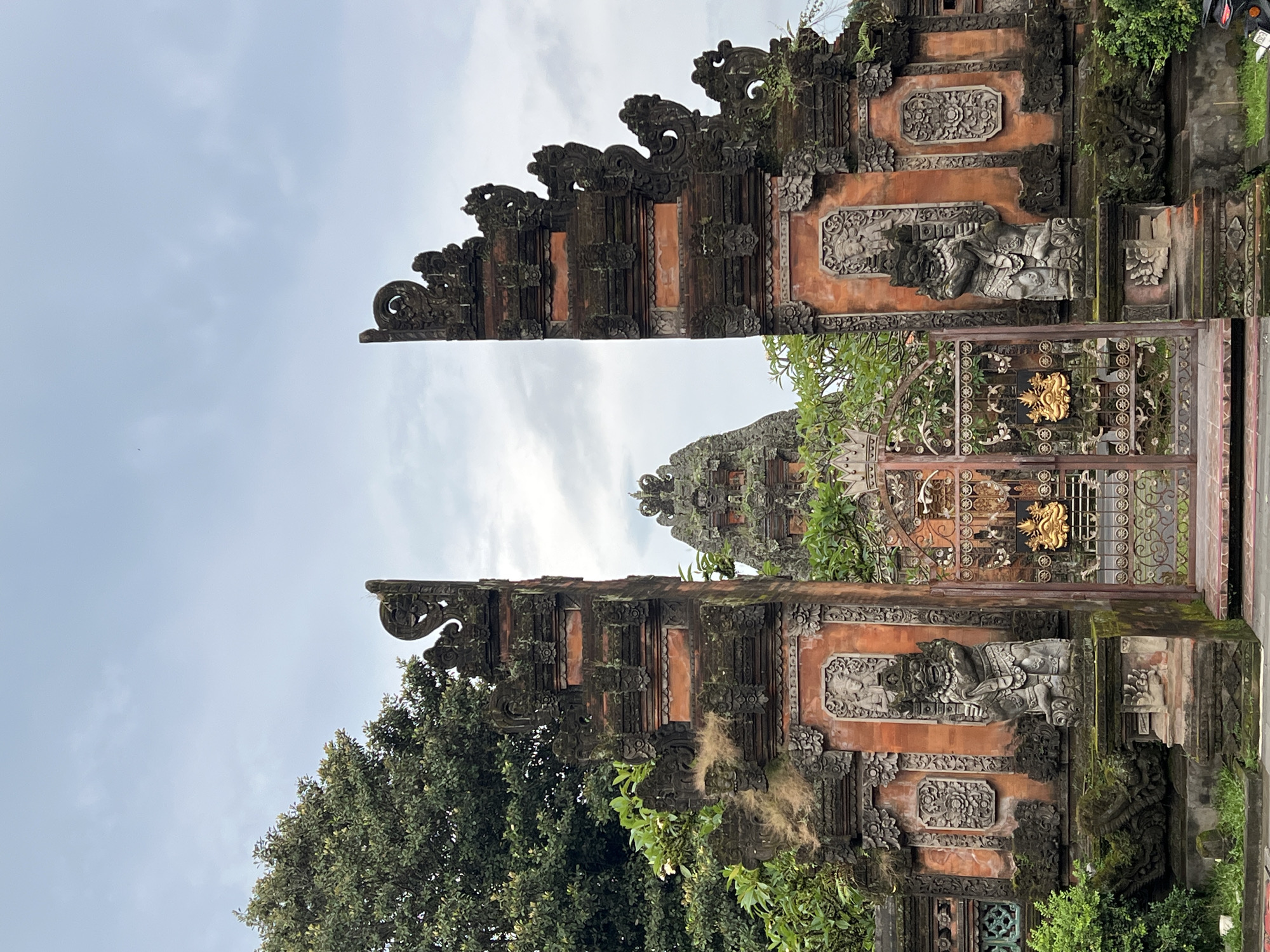 The image depicts a traditional Balinese gate in Peliatan, Indonesia. The gate consists of two ornately carved stone pillars flanking a metal gate with intricate gold accents. The pillars are decorated with carvings of mythical creatures and floral patterns, showcasing the region's artistic heritage. Lush greenery surrounds the gate, adding to the area's natural beauty and the structure's weathered appearance. The sky is overcast.