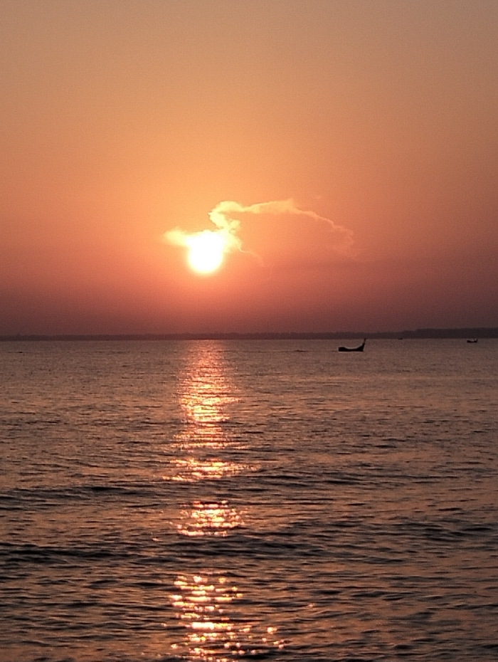 A sunset over water paints the sky in shades of orange and peach. The sun, partly obscured by a cloud, casts a shimmering reflection on the water's surface, creating a vertical stripe of light. Small boats are visible in the distance on the horizon. The water's surface is gently rippled, contributing to the sparkling effect of the reflected sunlight.