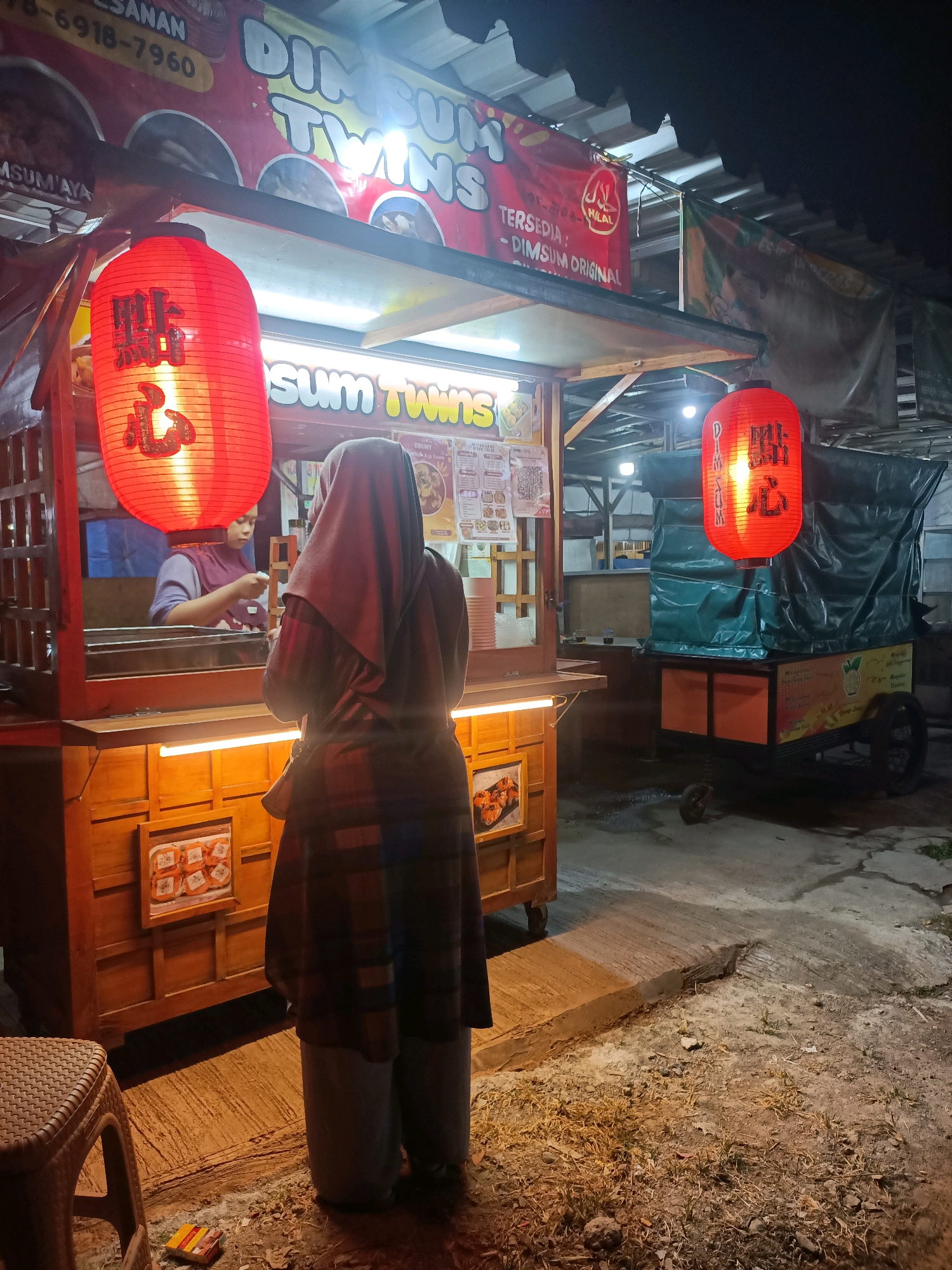 A woman wearing a dark hijab and a plaid outer garment stands facing a food stall at night. The stall, branded "DIM SUM TWINS," has a wooden front with an illuminated menu display. A woman wearing a purple top and a hijab is visible behind the counter, preparing food. Two red paper lanterns hang from the stall, one with Chinese characters. To the right, a covered cart is parked. The scene appears to be on a street in Wanajaya, Indonesia, with a paved surface and surrounding ground covered in dirt and dry grass.