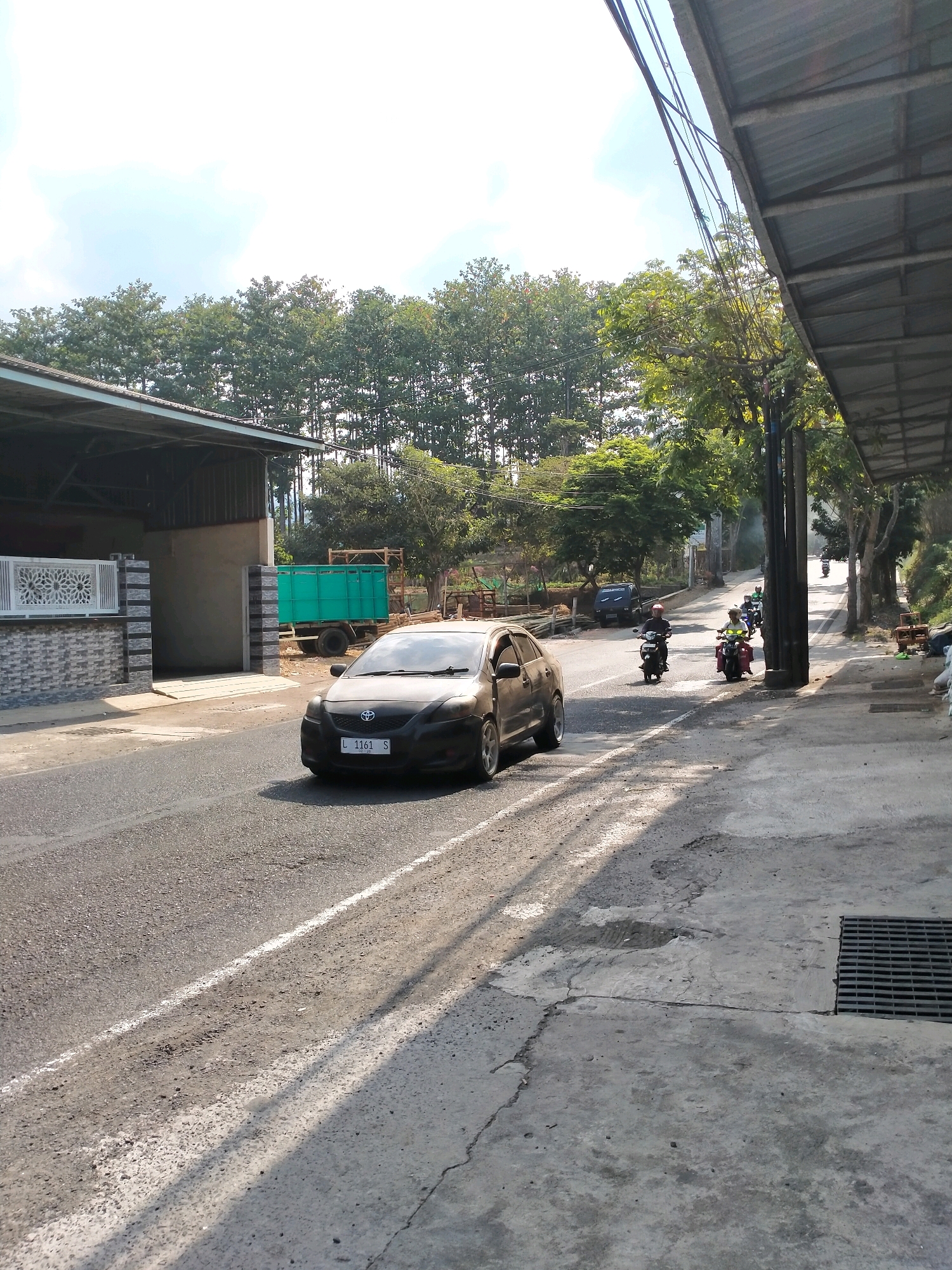 The image depicts a street scene in the city of Batu, Indonesia. A dark-colored sedan with license plate "L 1161 S" is driving on the right side of the road. The road has a white line marking the lane. On the left side of the road, there is a building with a gray and white patterned wall and a white decorative gate. Further down the road, a green truck is parked. Several people are riding motorcycles on the right side of the road, some of them in a convoy. The environment is characterized by a tree-lined street with a clear sky and some clouds. A metal awning extends from a structure on the right side of the road, and a storm drain is visible on the pavement. The lighting suggests it is daytime, with shadows cast by the trees and the awning.