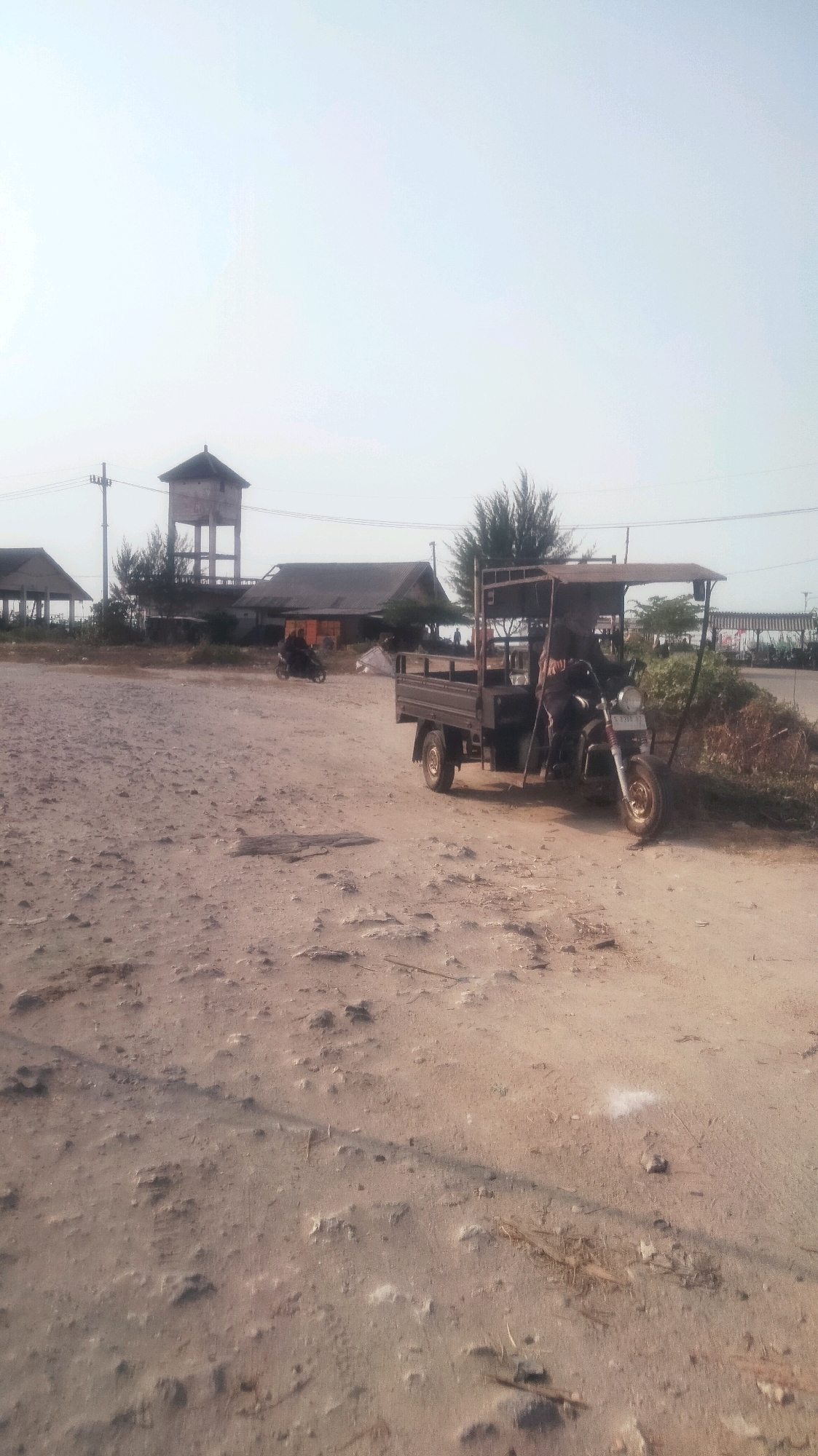 The image depicts an outdoor scene on a bright, overcast day, likely in Palang, Indonesia. The foreground is dominated by a sandy, uneven terrain with scattered debris and small rocks. In the midground, a three-wheeled motorcycle with a covered cargo bed and a driver is parked on a dirt path. The motorcycle has a license plate that appears to read "L 2518 KZ". Behind the motorcycle, there are several low-lying buildings with dark roofs, including one with a water tower structure behind it. Trees, including what appear to be pine trees, are visible between the buildings. Further in the background, there is a suggestion of a road with some sparse vegetation and what might be a distant structure or fence. Another motorcycle is visible in the distance, to the left of the three-wheeled vehicle, with a rider on it. The overall environment suggests a somewhat undeveloped or industrial area.