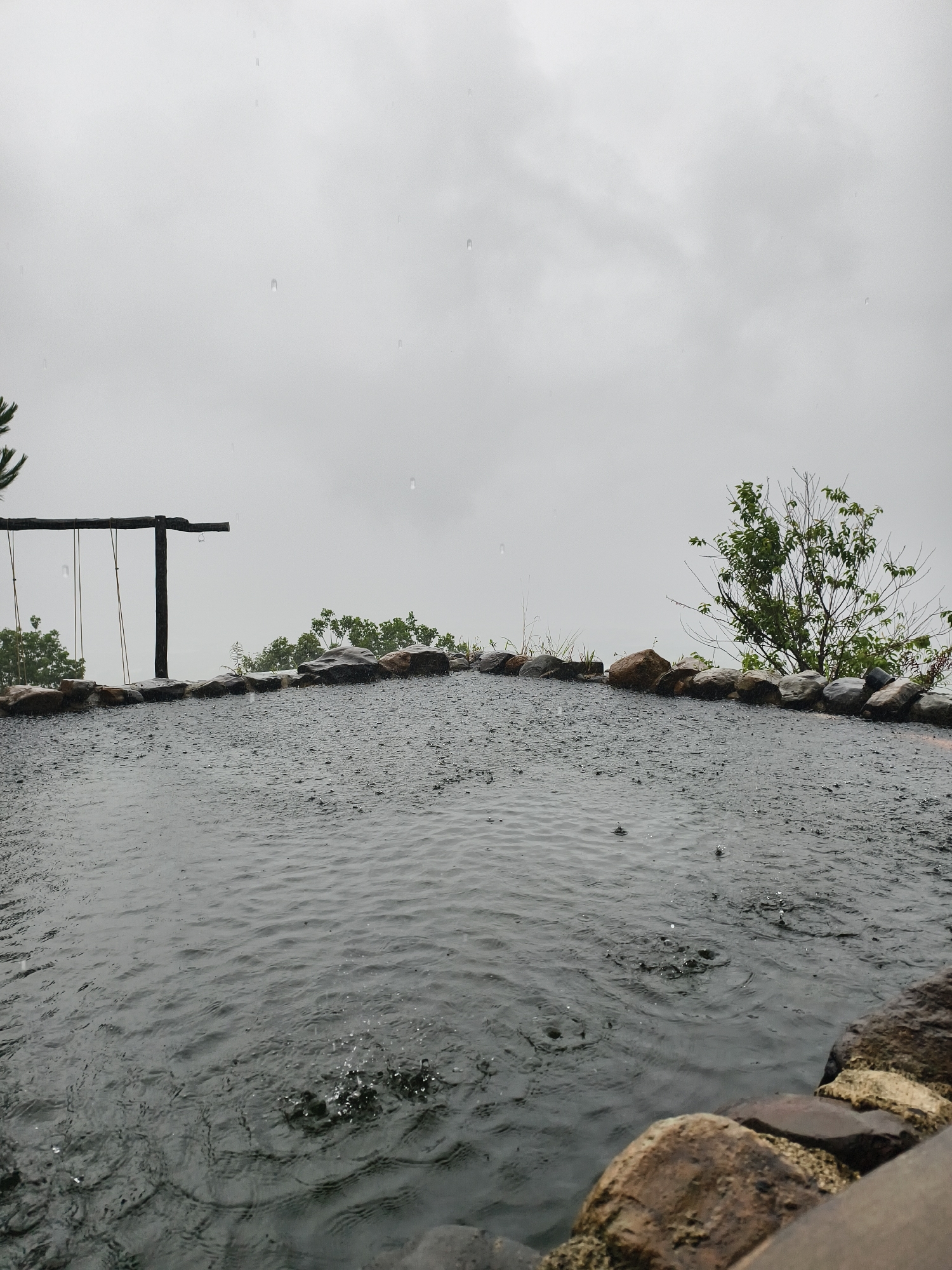 A landscape photograph taken on a rainy day shows a pool of water surrounded by rocks, trees, and a wooden structure. The sky is overcast and gray. The pool of water appears dark and rippled due to the raindrops hitting its surface. The surrounding rocks are also wet and dark. On the left side of the pool is a wooden structure resembling a swing set. Trees and other foliage are visible in the background. The overall setting is natural. The location is Unknown, Unknown.