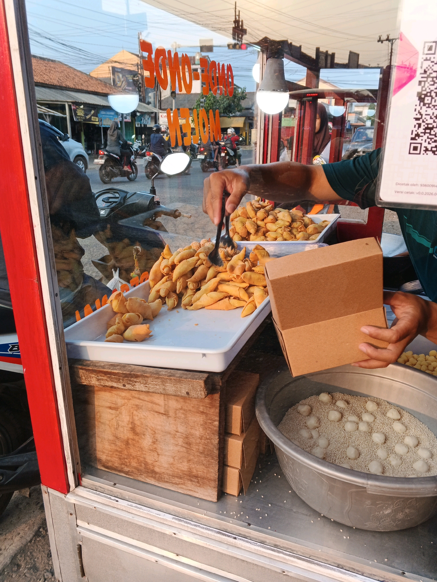 In Cilacap, Indonesia, a street vendor sells fried snacks through a glass-fronted stall. The stall displays a large tray of golden-brown, crescent-shaped fried items, possibly pastries or empanadas, and a smaller tray of similar items. In a large metal basin, white balls are being rolled in sesame seeds, with more seeds scattered around the basin. A person's hands are visible: one is holding a brown cardboard box, and the other is using a black utensil to scoop the fried snacks. The background seen through the glass shows a street scene with motorcycles, cars, buildings with tiled roofs, and overhead wires. A QR code is displayed on a pink and white flyer on the right side of the frame. The lighting suggests it is daytime, with the sun casting shadows and illuminating the scene.