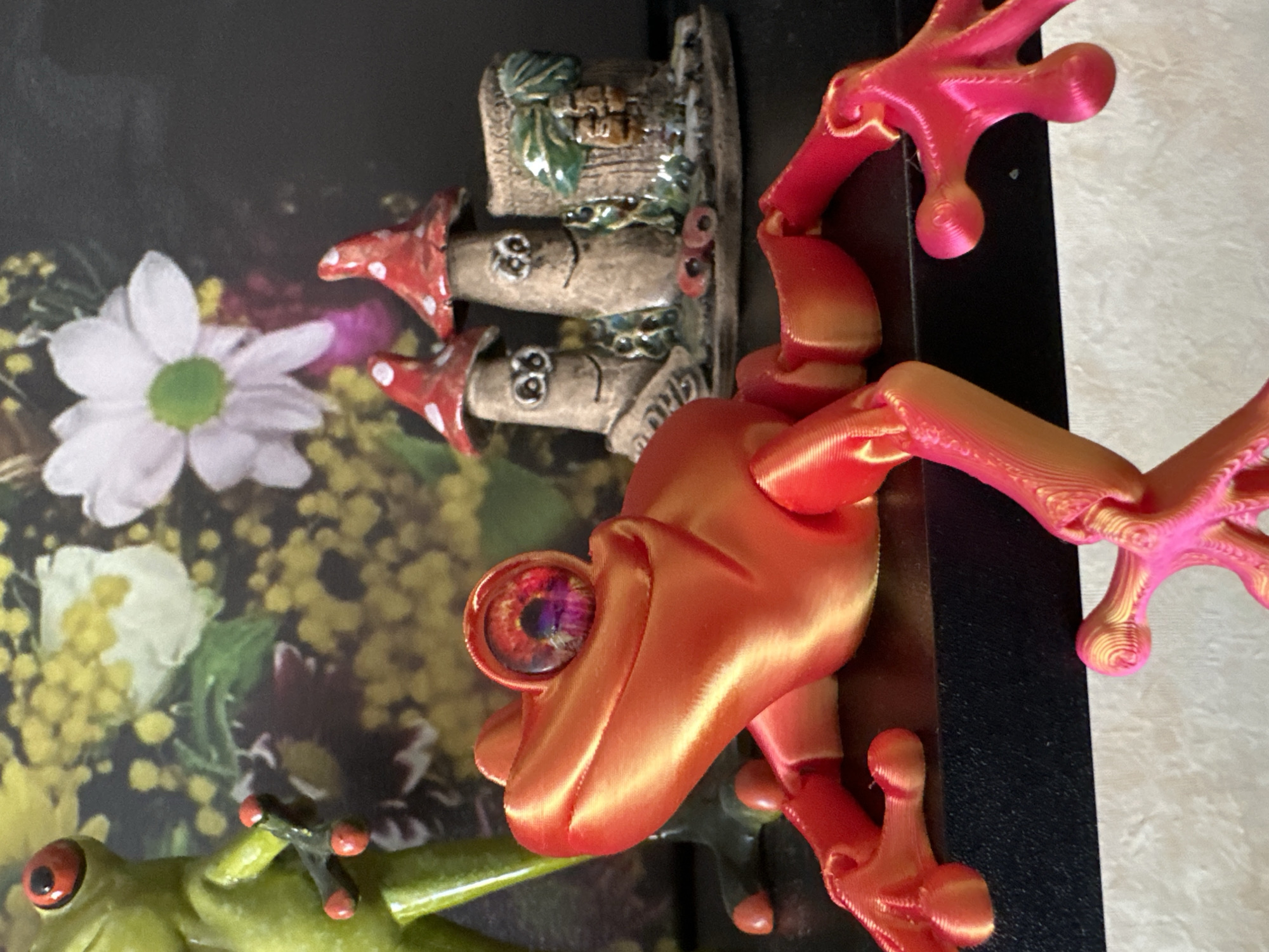 A close-up, eye-level shot captures a collection of decorative items, including several frog figurines and mushroom-shaped ornaments. In the foreground, an orange and pink 3D-printed frog lies on its stomach, its body and limbs articulated. Its large, multi-colored eye is the focal point. To the left and slightly behind, a green frog figurine stands upright. Behind the frogs, a cluster of three small, whimsical mushroom figures with faces are arranged. One mushroom has a hollowed-out log next to it. The background is a blurry depiction of flowers, with yellows, whites, and hints of pink. The objects are arranged on a black surface, which appears to be a shelf, with a textured, light-colored surface below. The scene is indoors, under artificial lighting. There is no indication of location in Podolsk, Russia.