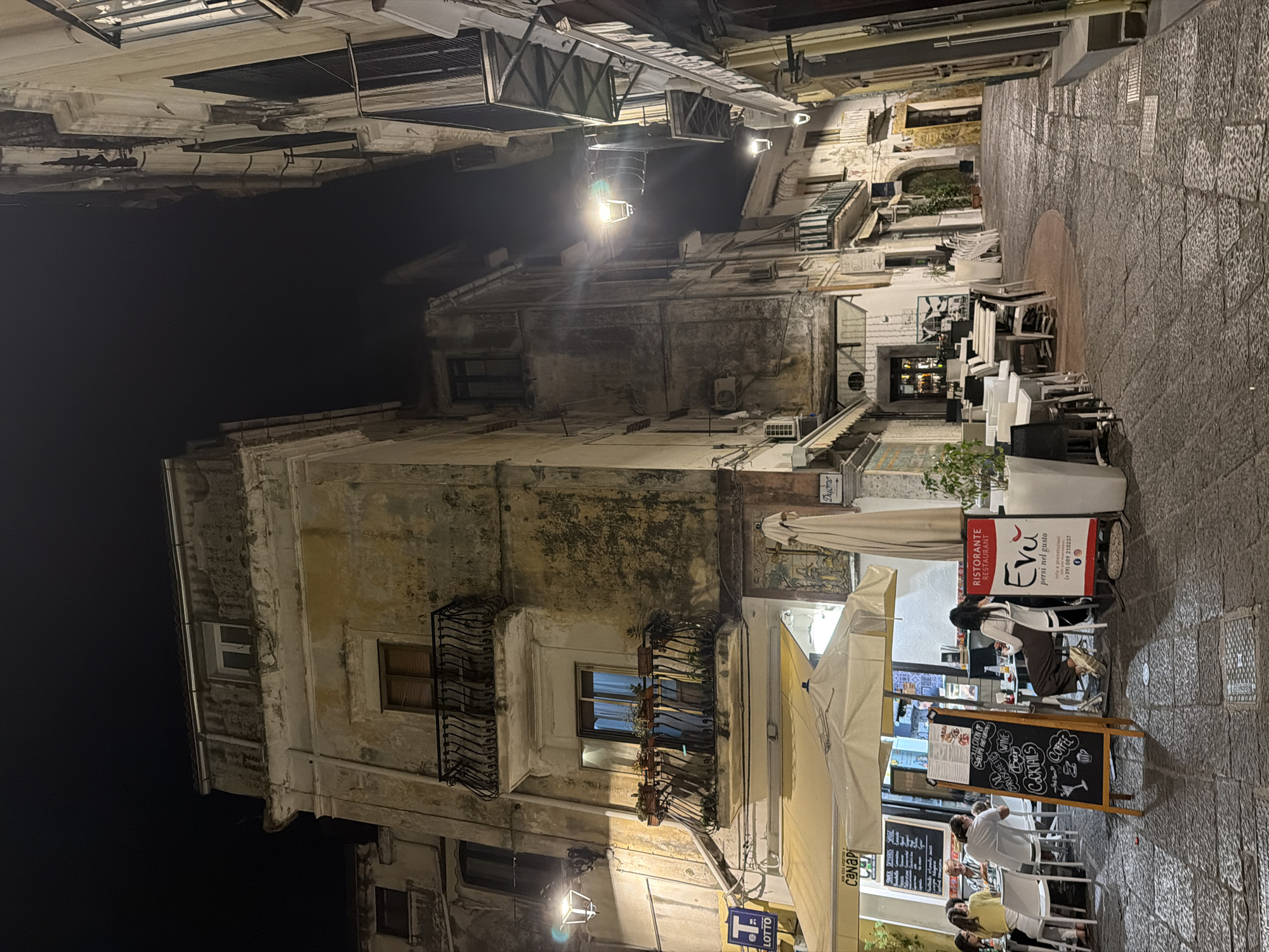 The image captures a vibrant night scene in an illuminated alleyway, characteristic of Vietri sul Mare, Italy. Old, multi-story buildings with weathered, light-colored facades line the street, many featuring traditional wrought-iron balconies adorned with potted plants. The sky is dark, indicating nighttime, with warm artificial light emanating from streetlamps, building interiors, and outdoor dining areas.
A prominent outdoor restaurant or café occupies the right side of the street (when viewing the image rotated correctly). White tables and chairs are arranged beneath large awnings and umbrellas, some filled with patrons. Several individuals are seated, socializing, or dining; a woman with dark hair is notably seated at a table near a large red and white sign.
The ground is paved with textured, possibly cobblestone, material. Various signs are visible, adding to the local charm. A large A-frame menu board displays offerings such as "COCKTAILS," "COFFEE," "WINE," and "SAULION OF BRUSCHETTE." The restaurant is identified by a sign reading "RISTORANTE Evu" with the tagline "persi nel gusto," along with contact information including an Italian phone number "(+39) 089 210237." Other visible text includes "LOTTO" and a sign pointing towards "Duomo," suggesting a nearby cathedral. The atmosphere appears relaxed and inviting, typical of an evening in a bustling Italian town.