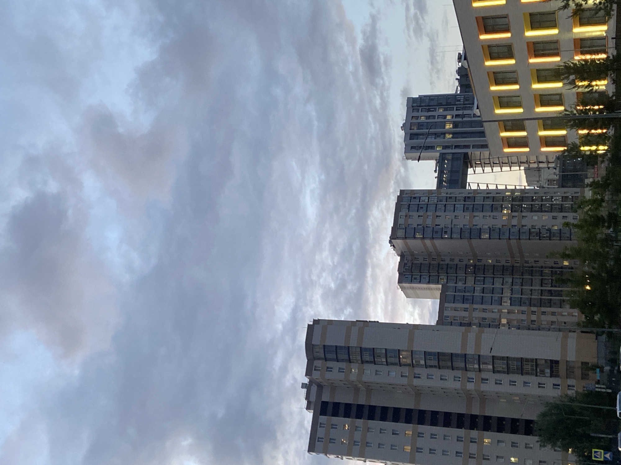The image captures a modern urban scene dominated by several tall residential buildings set against a cloudy sky. The perspective is angled slightly upwards, showcasing the towering structures and the atmospheric conditions above.
In the mid-ground and right side, multiple high-rise apartment buildings are visible. One prominent building on the far right features a striking facade with numerous square, yellow-orange illuminated sections, likely decorative elements or balconies with internal lighting, creating a warm glow. Other buildings are primarily constructed with grey and white panels, interspersed with dark windows. Many windows across the buildings are lit from within, indicating that the time of day is likely dusk or early evening.
The sky, occupying the upper and left portions of the image, is filled with soft, diffused clouds. These clouds range in color from light blue-grey to darker, more prominent grey patches, suggesting an overcast evening or fading daylight. The natural light is subdued, contrasting with the artificial illumination emanating from the buildings.
Towards the bottom right, green tree foliage partially obscures the base of the buildings. Subtle elements of a street scene are also visible, including a small portion of a white vehicle and two road signs. One sign is a yellow diamond with a black arrow, and another is a white rectangle with a red border and a diagonal red line, commonly denoting a "no stopping" or "no parking" restriction.
The scene depicts a static cityscape, devoid of people or overt activity. The overall impression is one of contemporary urban architecture under a transitional sky. No specific text is clearly legible on the buildings or signs beyond the basic shapes and colors of the traffic signs.