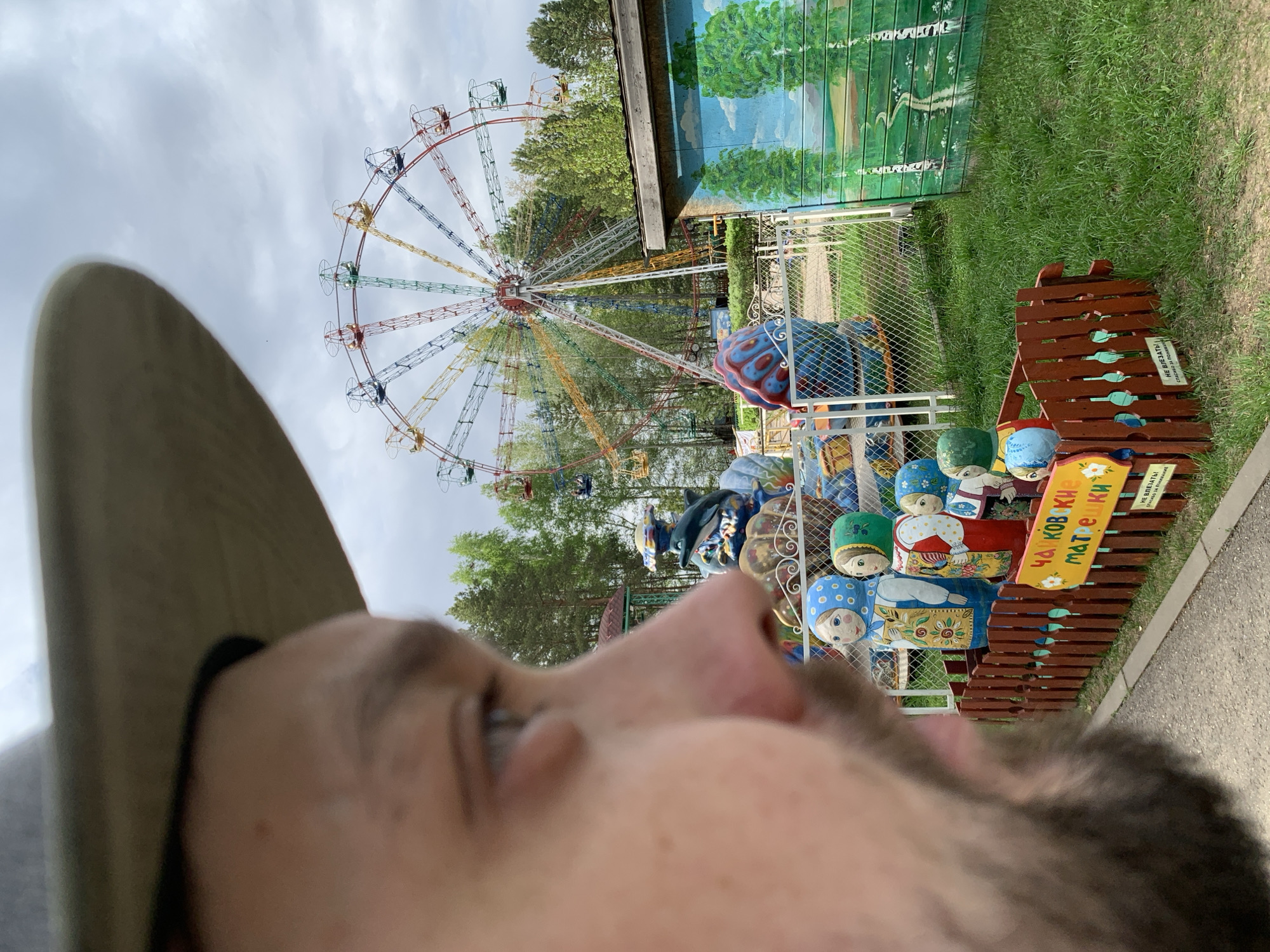A close-up, upward-angle shot shows a man in the foreground wearing a hat, with his face partially visible. In the background, a colorful Ferris wheel stands near a building decorated with a mural. In front of the building, there are painted wooden dolls and other decorative objects, possibly part of an amusement park attraction. Based on these clues and the question, the location is likely within the city of Chaikovsky, Russia.