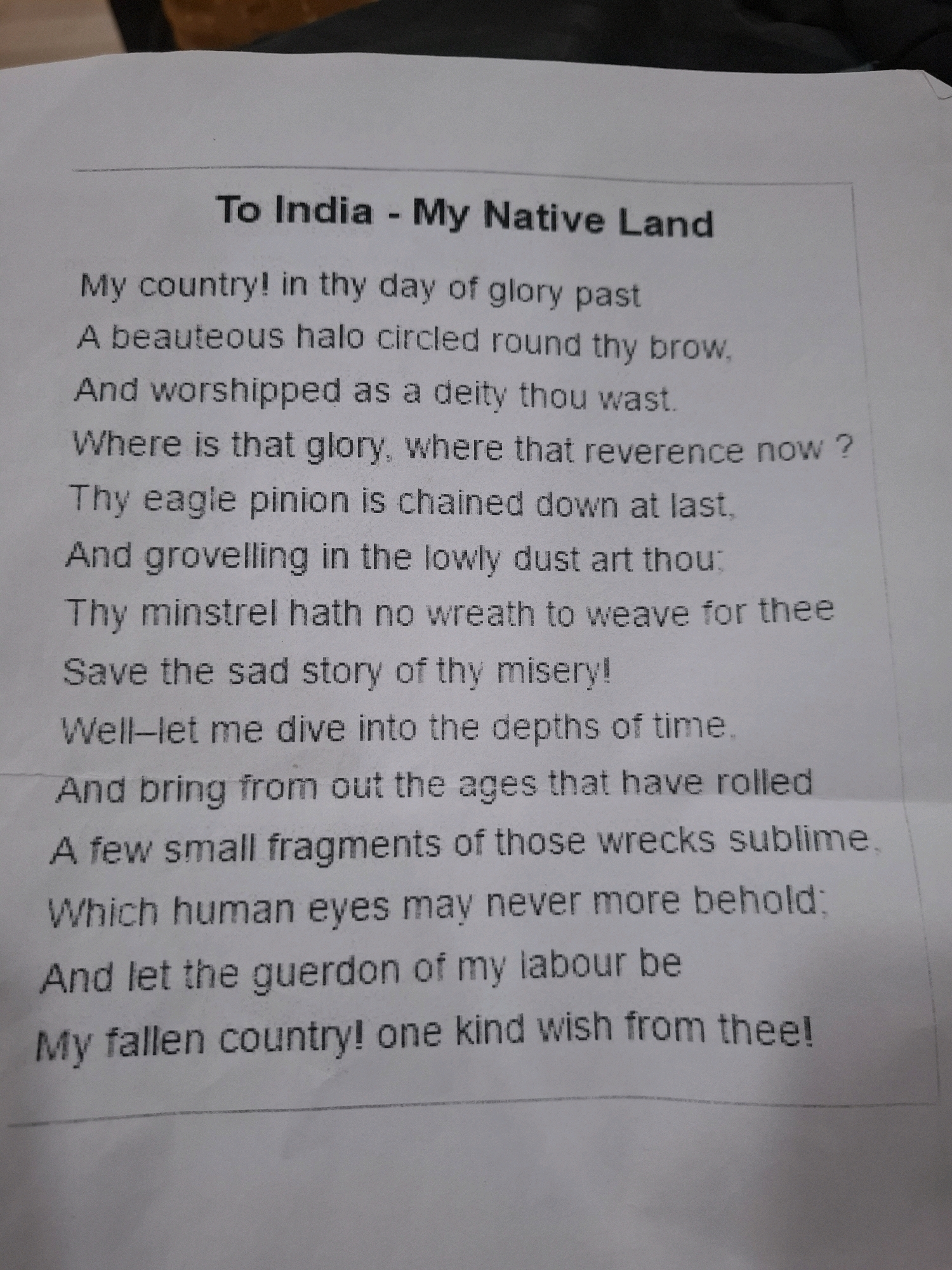 The image shows a close-up of a piece of paper with a poem titled "To India - My Native Land" printed on it. The poem is written in English and appears to be in a traditional poetic style. The paper is white and has some faint lines visible, possibly from a notebook or a lined sheet. The poem is centered on the page, with the title at the top. The text is clear and readable. The image is a still photograph and does not depict any specific location in Barasat, India, or any people or actions occurring in real-time. The background of the image is blurred and appears to be a surface, possibly a desk or table. There are no contextual clues within the image to suggest its origin or specific location beyond the content of the poem itself.