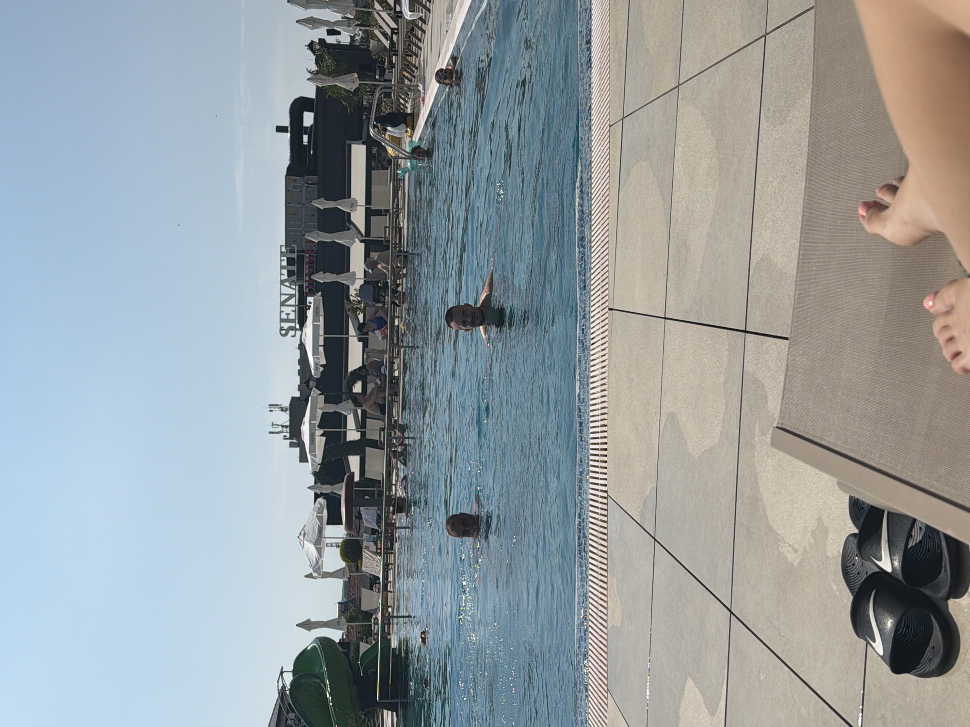 The image shows a sunny day at an outdoor swimming pool, likely at a hotel or resort. In the foreground, a person's feet, painted with pink nail polish, are resting on a lounge chair. A pair of black slides with white Nike logos are on the tiled ground next to the chair. The swimming pool is filled with blue water, and several people are swimming and relaxing in it. In the background, a green water slide is visible, along with white umbrellas and lounge chairs. A building with the word "SENATE" on it can be seen in the distance, suggesting the location may be related to a hotel named Senate. The overall atmosphere is one of leisure and recreation. The image is taken in Hisarya, Bulgaria, a city known for its mineral springs and spas.