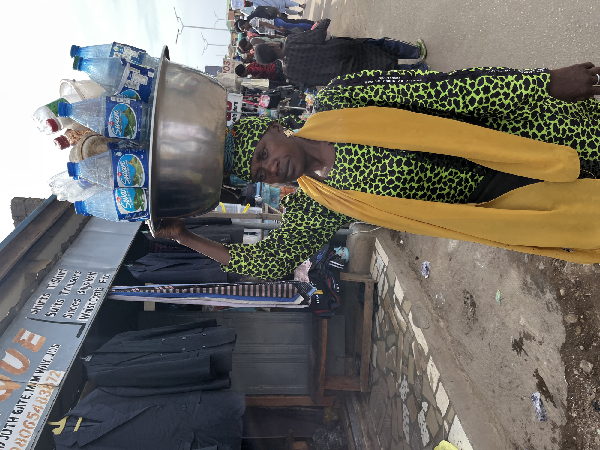 A street scene in Jos, Nigeria, features a woman carrying a large, silver-colored basin on her head, filled with multiple plastic bottles of "Swan Natural Spring Water." She is dressed in a vibrant green and black patterned outfit, complemented by a yellow shawl draped over her shoulders and a matching headwrap. Her expression is neutral as she balances her load.
In the background, other individuals are visible, moving about, suggesting a bustling market or commercial street environment. To the left, there is a shop or stall with a visible sign listing various clothing items for sale: "Shirts T-Shirt," "Suits Trousers," "Shoes Buys/Belt," and "Waist Coats Etc." The sign also includes a location, "JUTH GATE, M.M WAY, JOS," and a phone number, "08065483872." Other text on bottles includes "FUGUINÃO" and "RR-240604 PRINTED BY SUPER RR WAX."
The image appears to be taken during the daytime under clear skies. The ground is a mix of concrete pavement with some tiled sections and dirt. The scene captures a typical daily activity of a street vendor in an urban Nigerian setting.