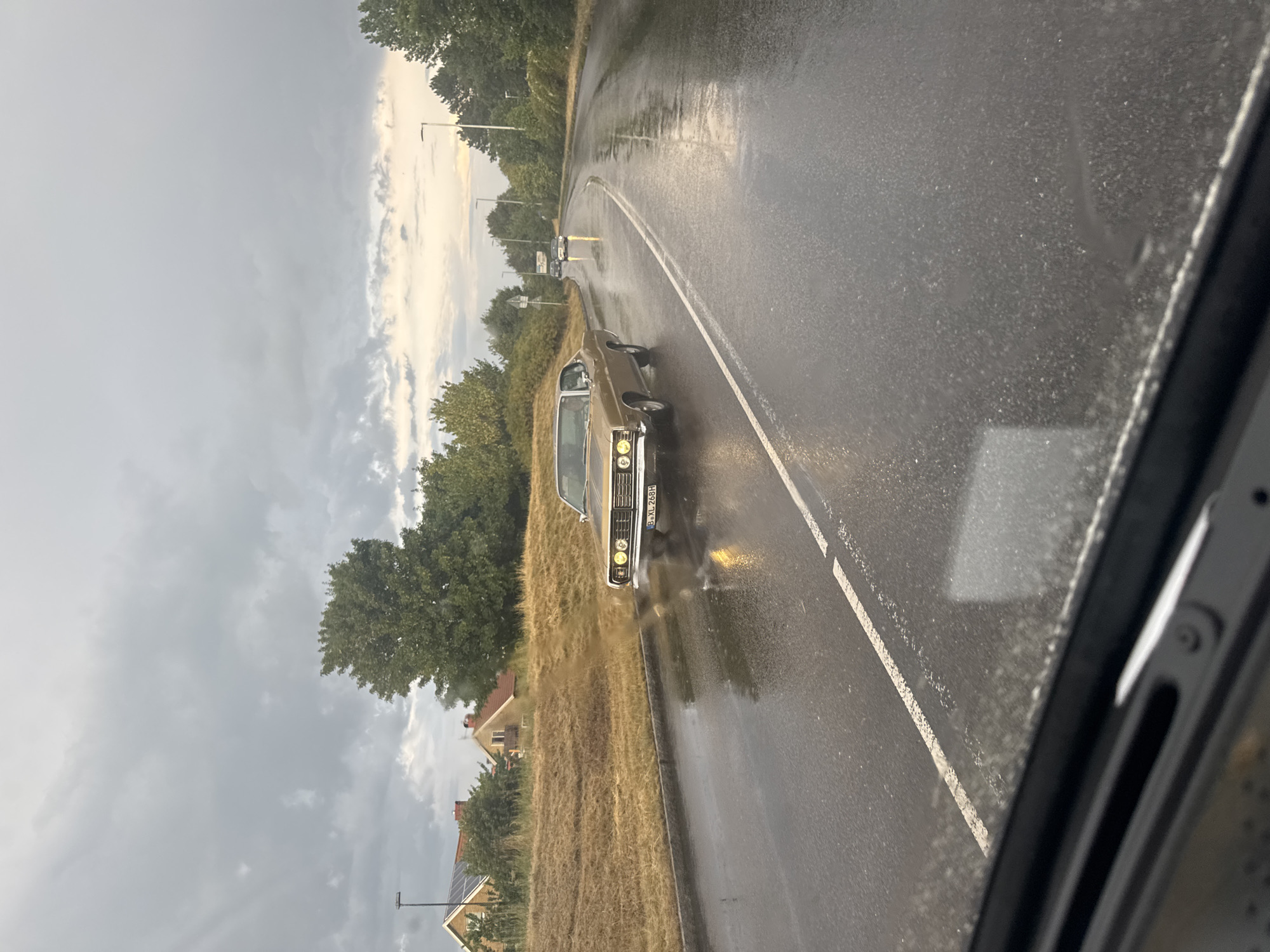 The image shows a gold-colored vintage American car driving on a wet road in Falkenberg, Sweden. The sky is overcast with dark clouds. The road is slick with rain, and reflections of streetlights and the car's headlights are visible on the surface. The car appears to be a Ford Galaxie from the late 1960s, identified by its distinctive grille and headlights. The license plate, partially visible, is B XL 268H.
On the left side of the road, there is a grassy embankment with some houses and trees in the background. On the right side of the road, there are more trees and streetlights. Another car is visible in the distance, also driving on the wet road. The perspective suggests the photo was taken from inside another vehicle. The overall scene depicts a gloomy, wet day in a suburban or semi-rural area of Falkenberg.