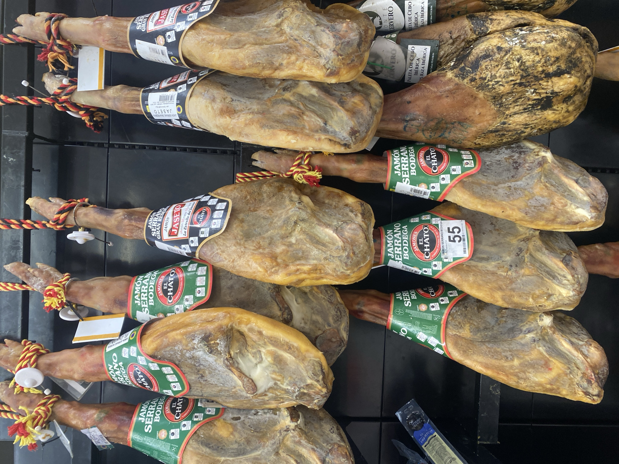 A supermarket display in Bel Air, Spain, features a row of cured hams, known as jamón serrano, hanging from hooks. Each ham is partially wrapped in green and red paper bands with Spanish text and branding. Some hams have yellow and red braided ropes attached to their tops. The background consists of a dark, tiled wall and shelving. The lighting is bright and appears to be overhead.