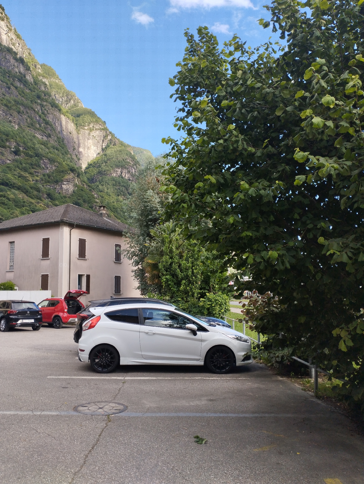A white Ford Fiesta is parked in a paved lot in front of a light brown building. The building has a dark brown roof and several windows with dark brown shutters. Behind the building are steep, tree-covered mountains. The sky is blue with a few white clouds. Several other cars are parked in the lot, including a red car with its trunk open and a black SUV. A large tree with green leaves is in the foreground, partially obscuring the view of the building and mountains. The image appears to have been taken on a sunny day.