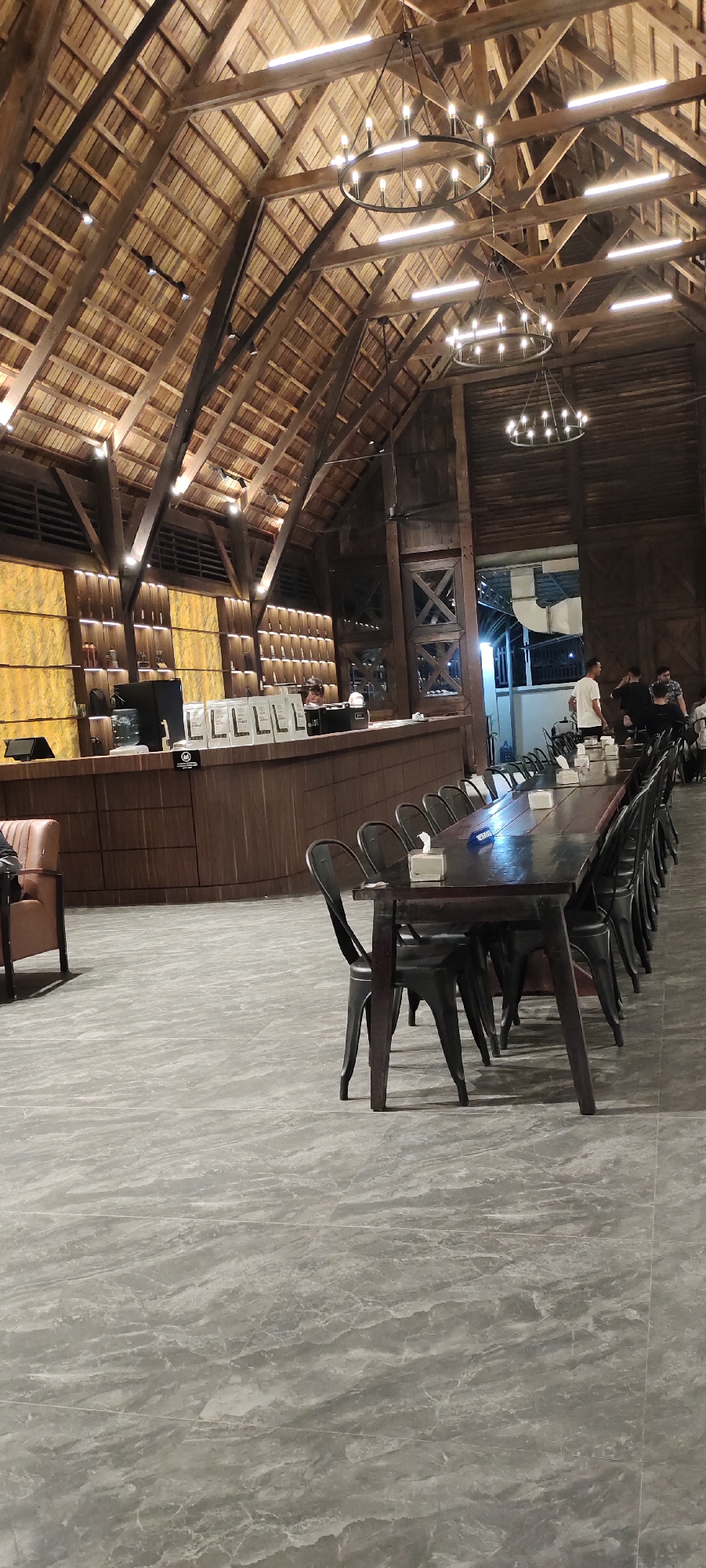 The image shows the interior of a spacious restaurant with a rustic and natural theme. The high, pitched ceiling is constructed with exposed wooden beams and a thatched roof, creating a warm and inviting atmosphere. Three large, circular chandeliers with multiple bulbs hang from the ceiling, providing ample lighting.
In the foreground, a long, dark wooden table is set with rows of black chairs. Tissues are placed on the table, suggesting it's ready for diners. The floor is covered with large, grey marble-patterned tiles.
In the background, a long wooden counter area appears to be a service bar or a point of sale. Several white bags, possibly containing coffee or other products, are neatly arranged on the counter. Behind the counter, shelves are stocked with various items, and a few people can be seen working or interacting. One person is leaning over the counter, possibly serving a customer. Further back, there's an open doorway leading to another area where more people are visible, some standing and chatting. The overall impression is that of a bustling restaurant during its operating hours, likely in the evening given the interior lighting. The design elements, particularly the wood and thatched roof, suggest a location that embraces natural materials and a somewhat rural or bohemian aesthetic. There are no explicit signs of weather or time of day beyond the interior lighting.