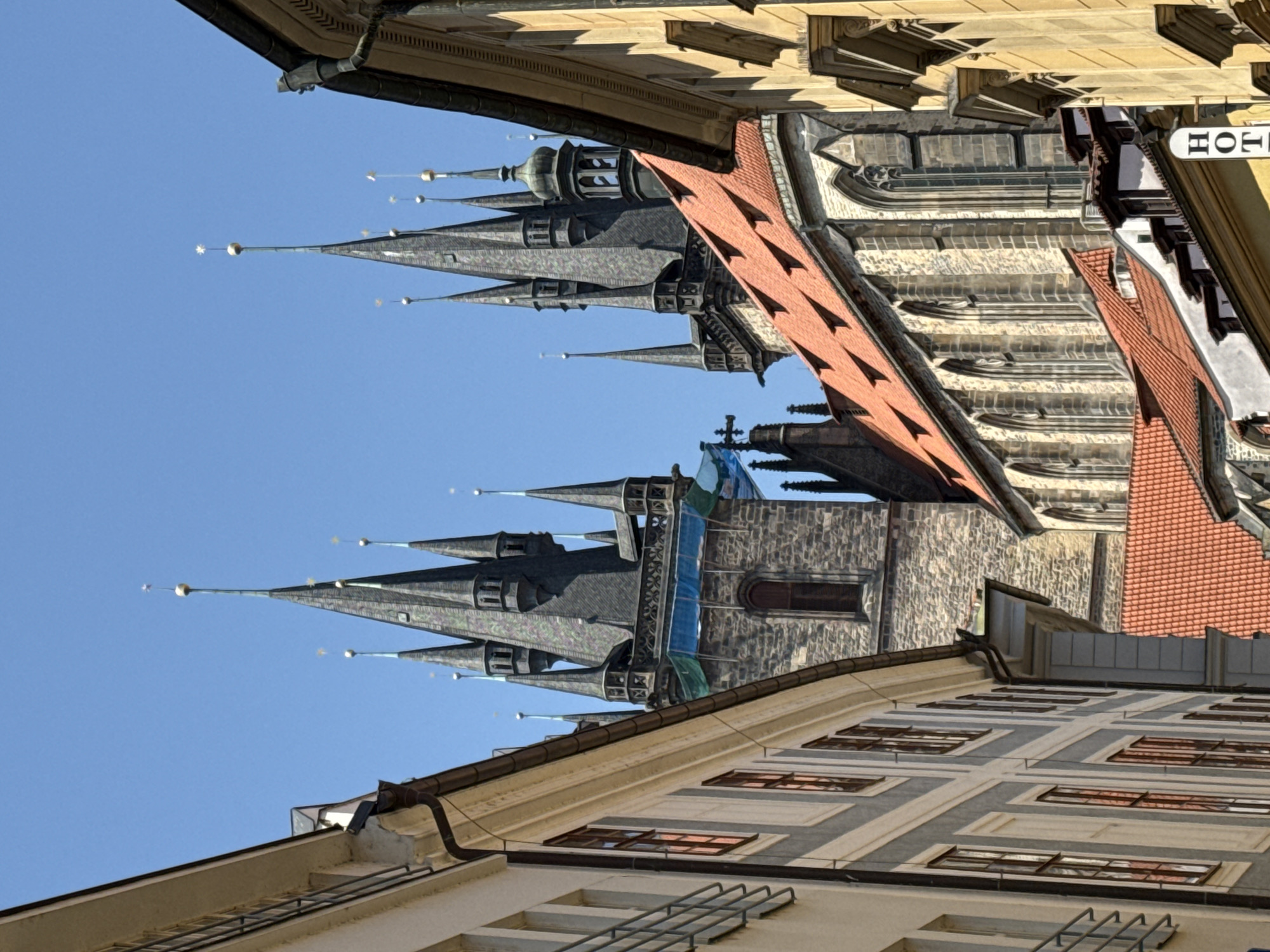 The image shows a view of the towers of the Church of Our Lady before Týn in Prague, Czechia. The church is situated in the Old Town Square and is a prominent landmark of the city. The photograph is taken from a narrow street, with buildings on either side framing the view of the Gothic towers. The sky is clear and blue. The architecture is characterized by its steep roofs, spires, and stone construction. The overall impression is one of historical grandeur and architectural magnificence.