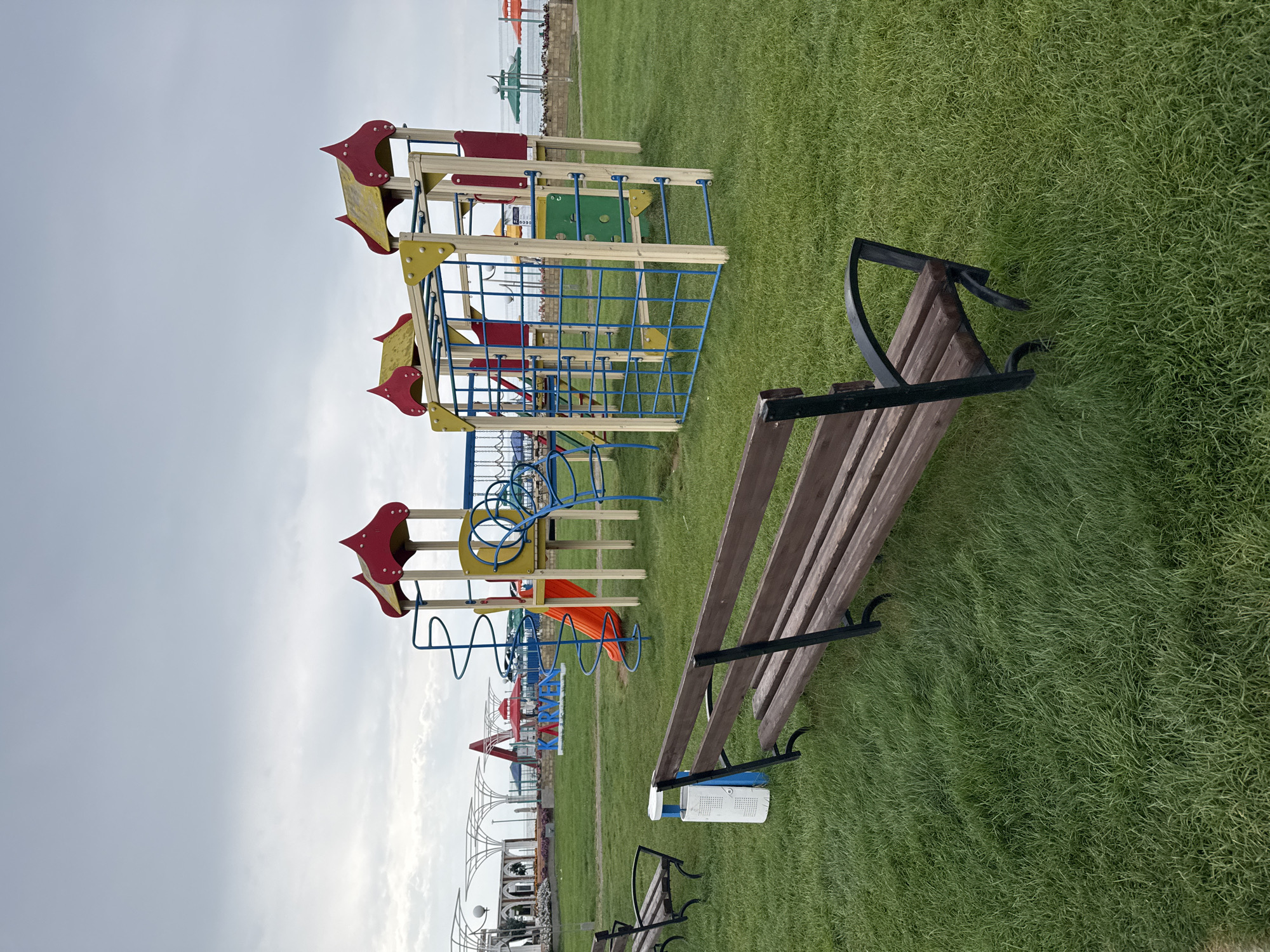 The image depicts an outdoor children's playground situated on a well-maintained green lawn in Sary-Oy, Kyrgyzstan. In the foreground, two dark wooden park benches with black metal frames are positioned facing away from the viewer, one closer and one further back. Adjacent to the closer bench is a blue and white trash receptacle. The playground equipment is brightly colored, featuring a central structure with red and yellow peaked roofs, a bright orange slide, blue spiral slides, and a blue climbing net. In the background, beyond the playground, a paved area is visible with decorative white metal structures, white light posts, and what appear to be small trees or bushes. Further in the distance, there are umbrellas, suggesting a proximity to a beach or resort area. The sky is overcast with a light grey cloud cover. There are no people visible in the scene.