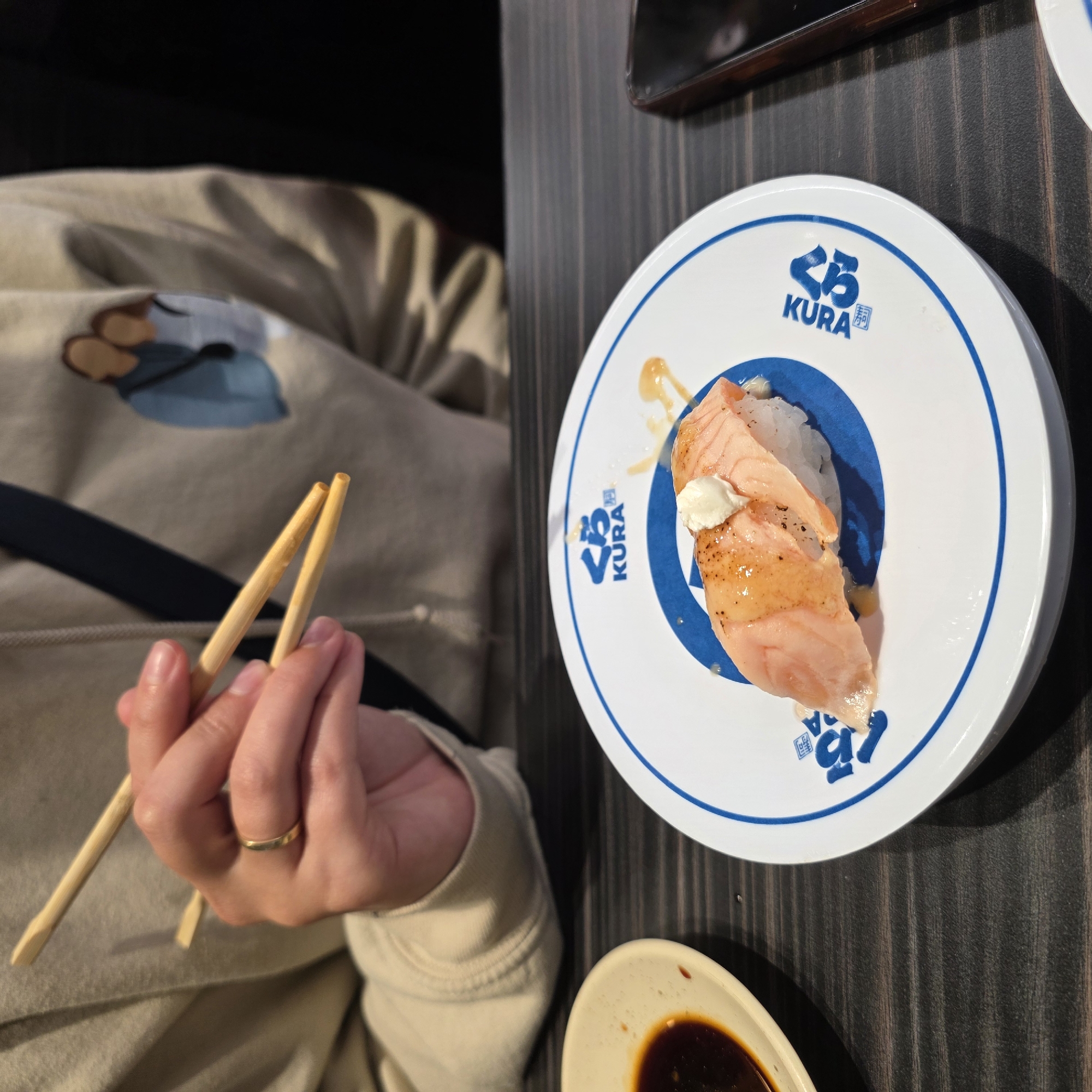 An overhead, medium shot captures a person's hands holding chopsticks over a plate of sushi at a table in a restaurant. The plate is white with a blue rim and features the Kura Sushi logo. A single piece of salmon nigiri sushi with a dollop of white sauce sits on the plate. To the left of the sushi plate is a small white dish containing dark soy sauce. The person's left hand, adorned with a gold wedding band, holds two wooden chopsticks, one of which is positioned to pick up the sushi. The background shows the person wearing a tan hoodie with a graphic design of two abstract figures on the left sleeve. The setting appears to be a Kura Sushi restaurant, common in Beaverton, Oregon, a suburb of Portland, Oregon.