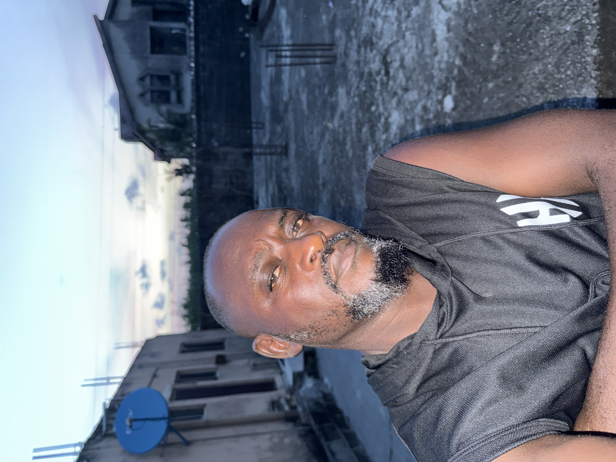 The image shows a man with dark skin, a bald head, and a salt-and-pepper beard, wearing a black athletic shirt. He is outdoors, likely on a rooftop in Ado, Nigeria, based on the surrounding urban landscape. Behind him are buildings of varying heights, some with visible satellite dishes, suggesting residential or commercial structures. The sky is visible in the background with subtle colors indicating dusk or dawn. The scene appears to be at a high angle, as the man is positioned looking downward towards the viewer.