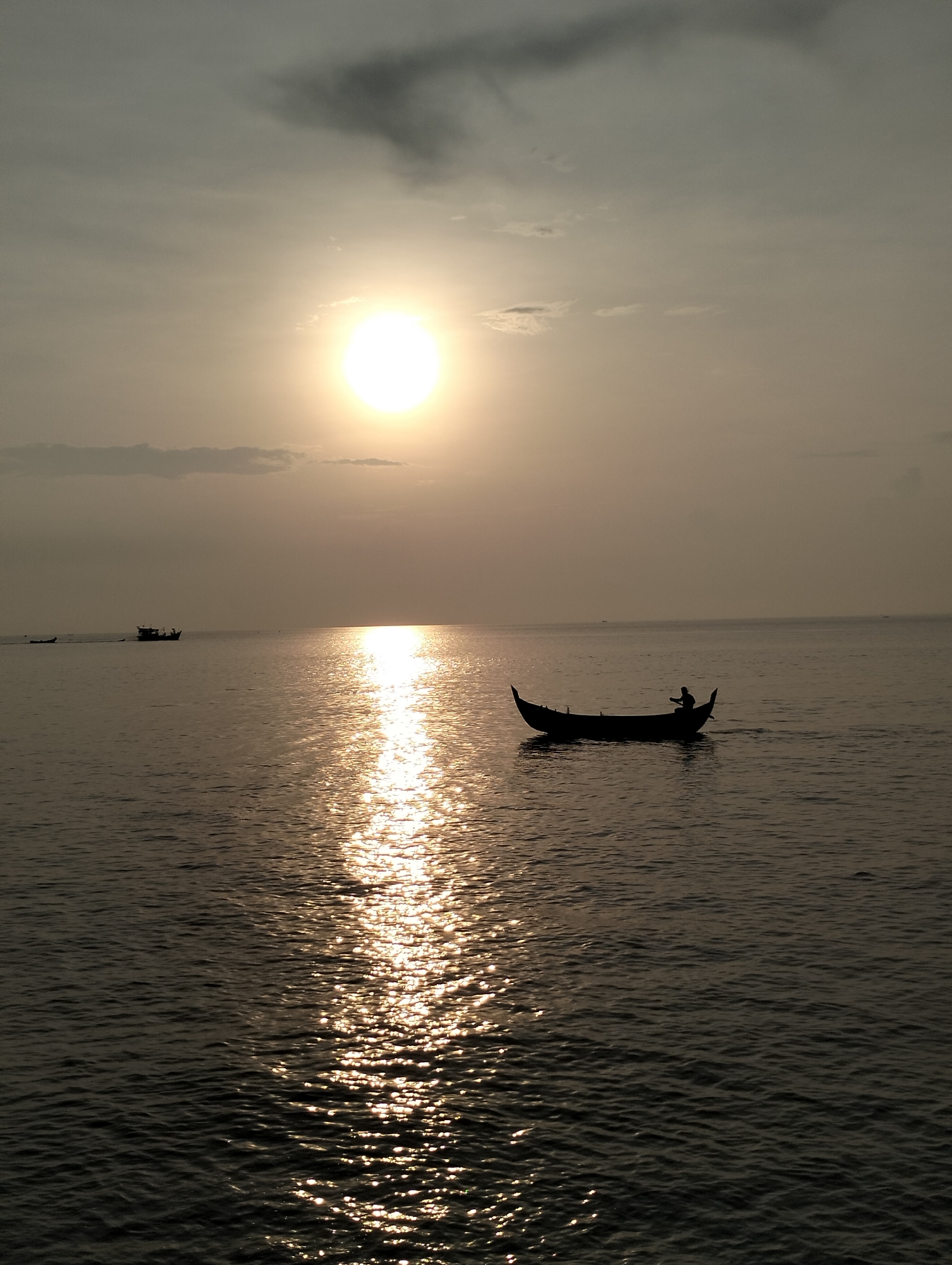 The image portrays a tranquil maritime scene at either sunrise or sunset, characterized by a bright, circular sun dominating the upper right portion of the frame. The sun's intense light creates a vibrant, shimmering path of golden reflections stretching across the calm expanse of water towards the viewer.
In the mid-ground, a small, traditional boat is silhouetted against the brilliant sunlight on the water. A lone person is seated within this boat, also appearing as a dark silhouette, seemingly navigating or attending to something. Further in the distance, on the left side of the frame, another larger boat, possibly a fishing vessel, is faintly visible, also silhouetted against the muted horizon. The water's surface exhibits gentle ripples, particularly pronounced where the sunlight reflects.
The sky above is a soft, diffused grey, with some scattered, darker clouds present in the upper left quadrant. The overall atmosphere evokes a sense of peace and natural beauty. The setting is likely a coastal area, specifically Lhokseumawe, Indonesia, given the context. There is no visible text in the image.