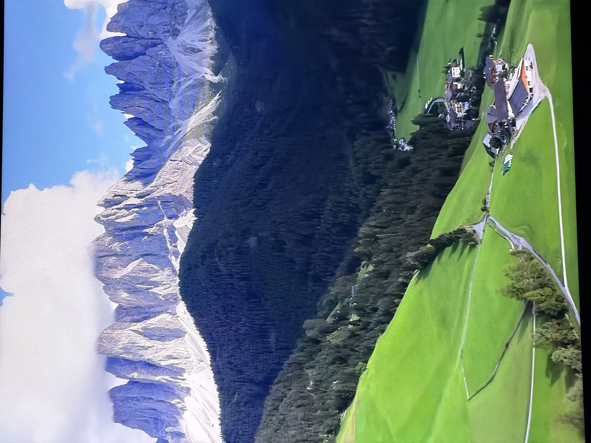 The image depicts a majestic mountain range with sharp, jagged peaks partially obscured by clouds. Below the mountains, a dense, dark green coniferous forest covers the slopes. In the foreground, vibrant green rolling hills lead down to a small village. The village consists of several buildings with traditional architecture, nestled amidst the trees and fields. A winding road connects the village to the surrounding landscape. The sky above is blue with scattered white clouds. The scene conveys a sense of natural beauty and tranquility. The location is not in the city of Chirchik, Uzbekistan, but rather appears to be in the Alps, given the distinctive mountain formations and architectural style of the village.