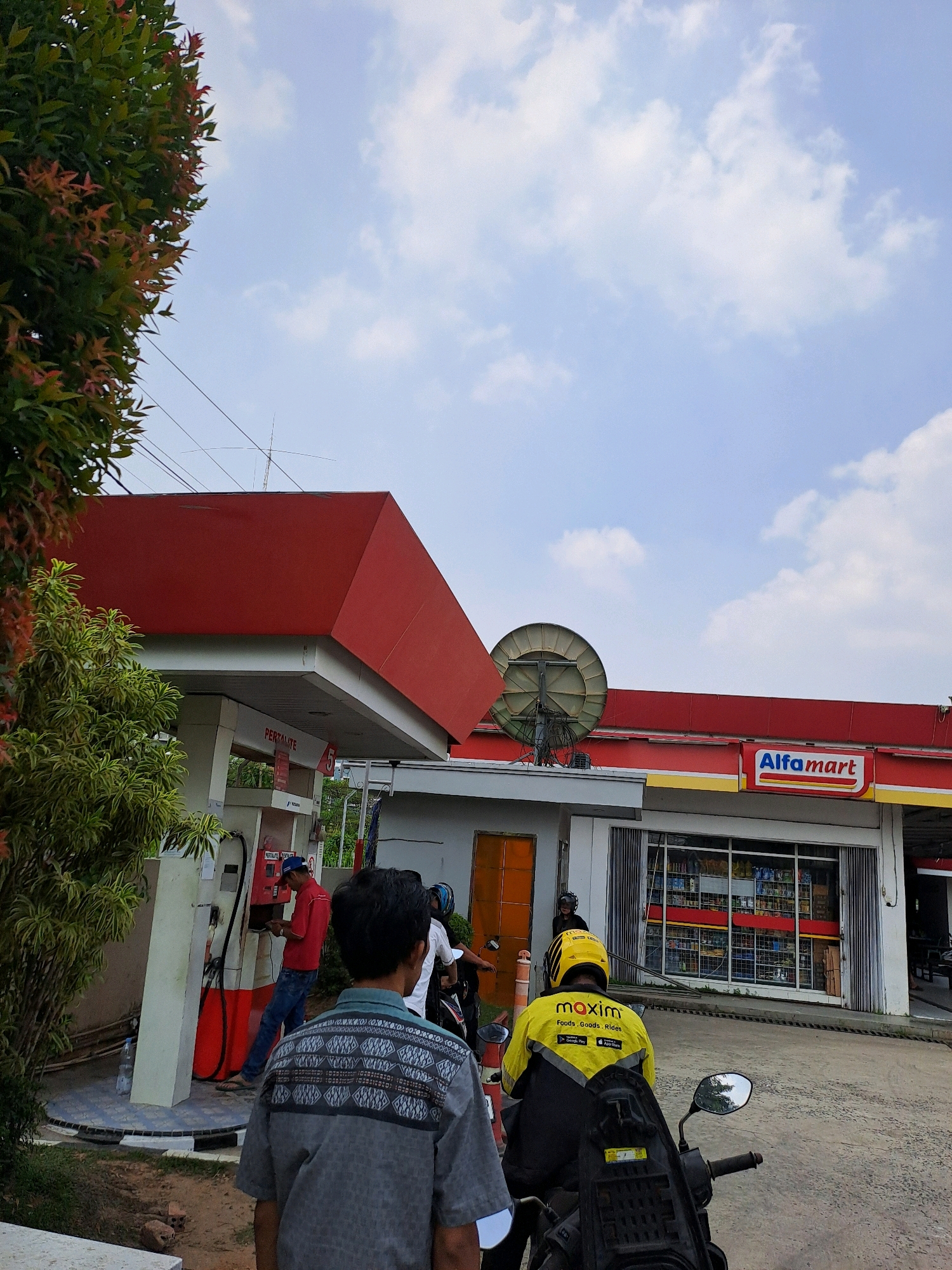 The image captures an outdoor scene at a gas station and convenience store. In the foreground, the back of a person wearing a gray patterned shirt is visible. To the left, a person in a red shirt and blue jeans is interacting with a gas pump. Behind them, a motorcycle with a rider wearing a yellow jacket and helmet, identified with the "maxim" logo, is parked. Further to the right, the facade of an "Alfamart" convenience store is visible, with shelves of products displayed through the windows. A large satellite dish is mounted on the roof above the store. The environment includes a red-roofed canopy over the gas pumps, a cloudy sky, and greenery from trees and bushes. The overall impression is that of a commercial area in Palembang, Indonesia, likely during daylight hours.
