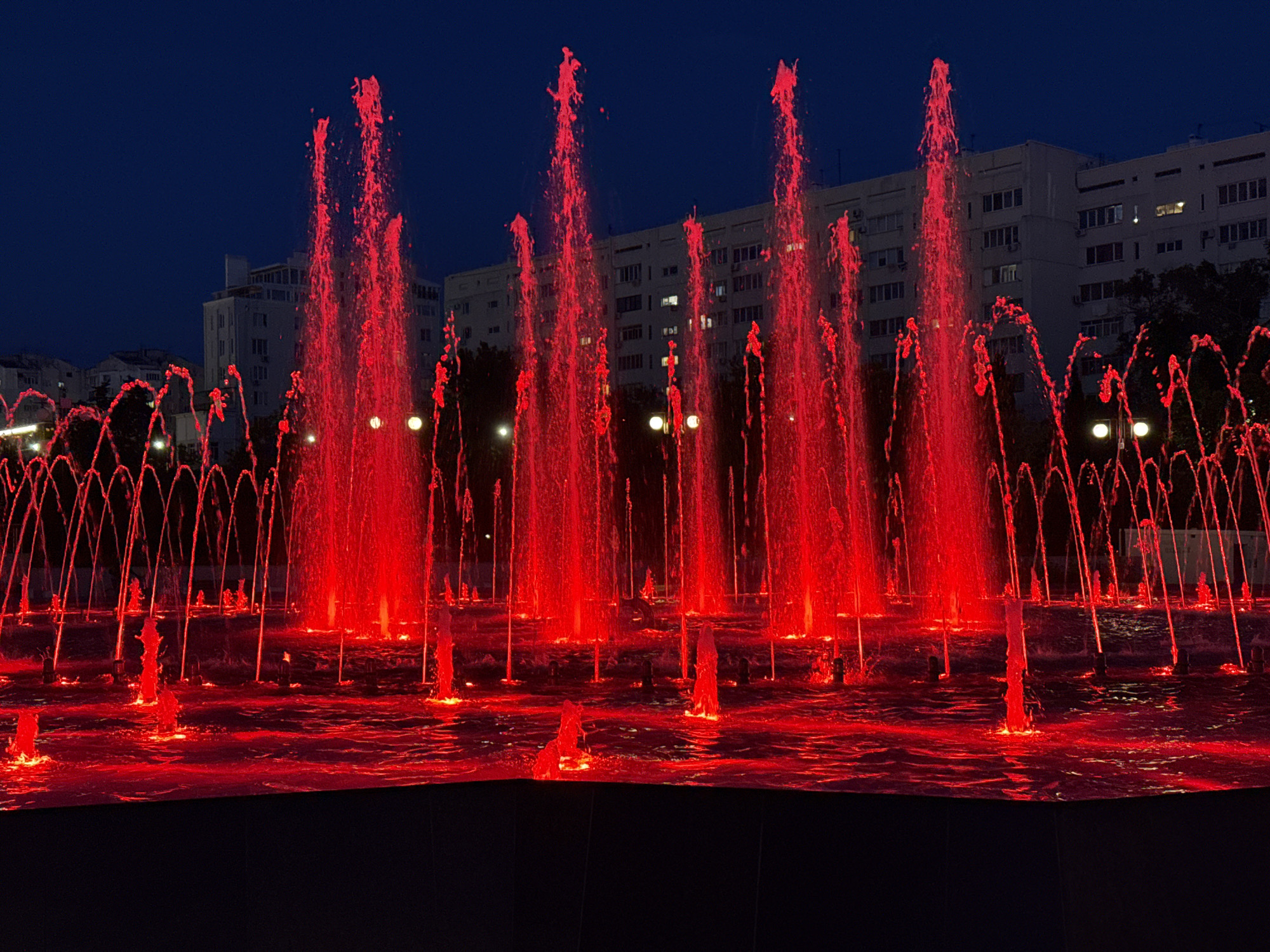 In an urban environment at dusk, a large water fountain is illuminated with vibrant red lights, casting a dramatic glow on the cascading water. The fountain features numerous jets of water shooting upwards, with some reaching significant heights, creating a dynamic and colorful display against the dark blue sky. In the background, multi-story buildings with visible windows are silhouetted, suggesting a city setting. The water in the fountain basin is also illuminated red, reflecting the bright lights. The scene appears to be a public space, possibly a park or plaza, designed for nighttime viewing and enjoyment.