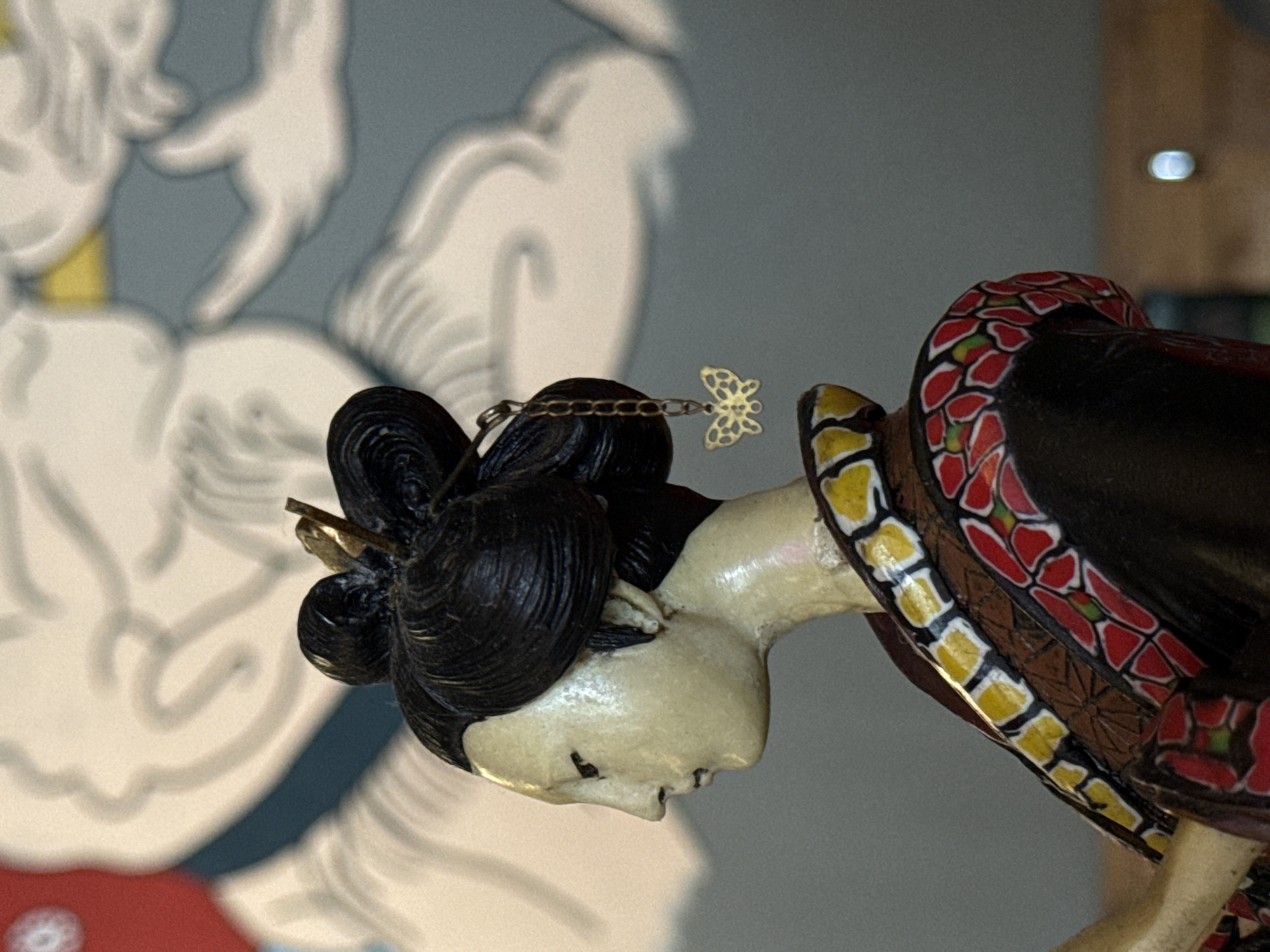 A close-up shot depicts a Japanese geisha figurine in a bowing pose. The figurine has pale skin, a black coiled hairstyle adorned with a gold-colored pin and a chain leading to a butterfly-shaped charm. The neckpiece is decorated with a pattern of red and green floral shapes outlined in white. The background shows a blurred mural in blues, whites, and reds. The location is Krasnoyarsk, Russia.
