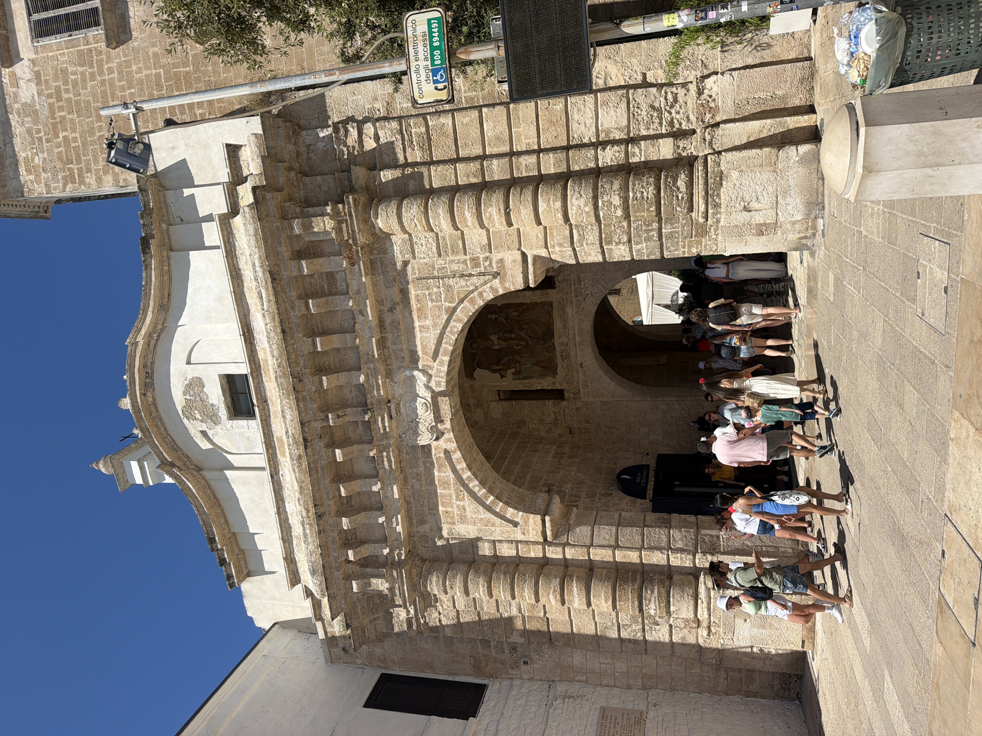 The image shows the Porta Vecchia, a historic stone archway in the city of Polignano a Mare, Italy. The archway, made of rough-hewn stone blocks, features classical architectural elements like fluted columns and decorative carvings. Above the arch, a white building with a rounded facade and a small dome is visible. Sunlight casts strong shadows, indicating a bright, sunny day with a clear blue sky.
In front of the archway, a group of people are walking and standing. They are dressed in casual summer clothing. Some are entering or exiting the archway, suggesting it's a thoroughfare. A sign with Italian text and a wheelchair symbol is mounted on a pole to the right of the arch, indicating controlled access and an emergency number. Further to the right, a large, dark digital display board is attached to the pole. A green trash receptacle overflowing with plastic bottles and bags is situated at the bottom right corner of the frame. The ground is paved with stone slabs.