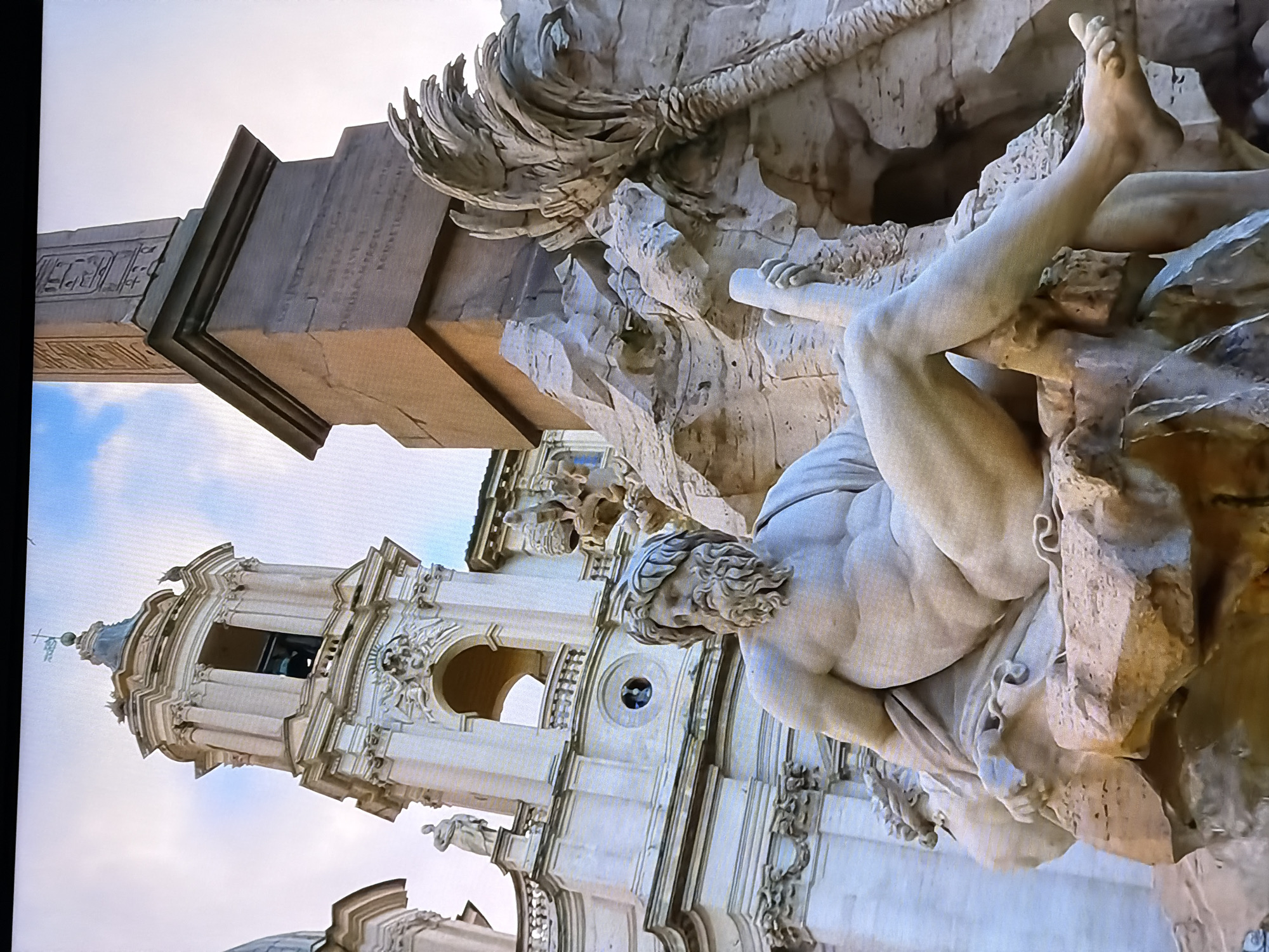 The image shows a section of the Fountain of Four Rivers in Piazza Navona, Rome, Italy. The fountain, designed by Gian Lorenzo Bernini, depicts four river gods representing the Nile, Ganges, Danube, and Río de la Plata. In the foreground, a male figure, sculpted from marble, reclines on rocks. The figure has a beard, a laurel wreath on his head, and appears muscular. He is holding a staff and water flows from the rocks around him, suggesting a river. Behind the fountain, an ornate building with a bell tower and a tall obelisk are visible. The sky is partly cloudy. The prompt incorrectly places this landmark in Chirchik, Uzbekistan; this is a well-known landmark in Rome, Italy.