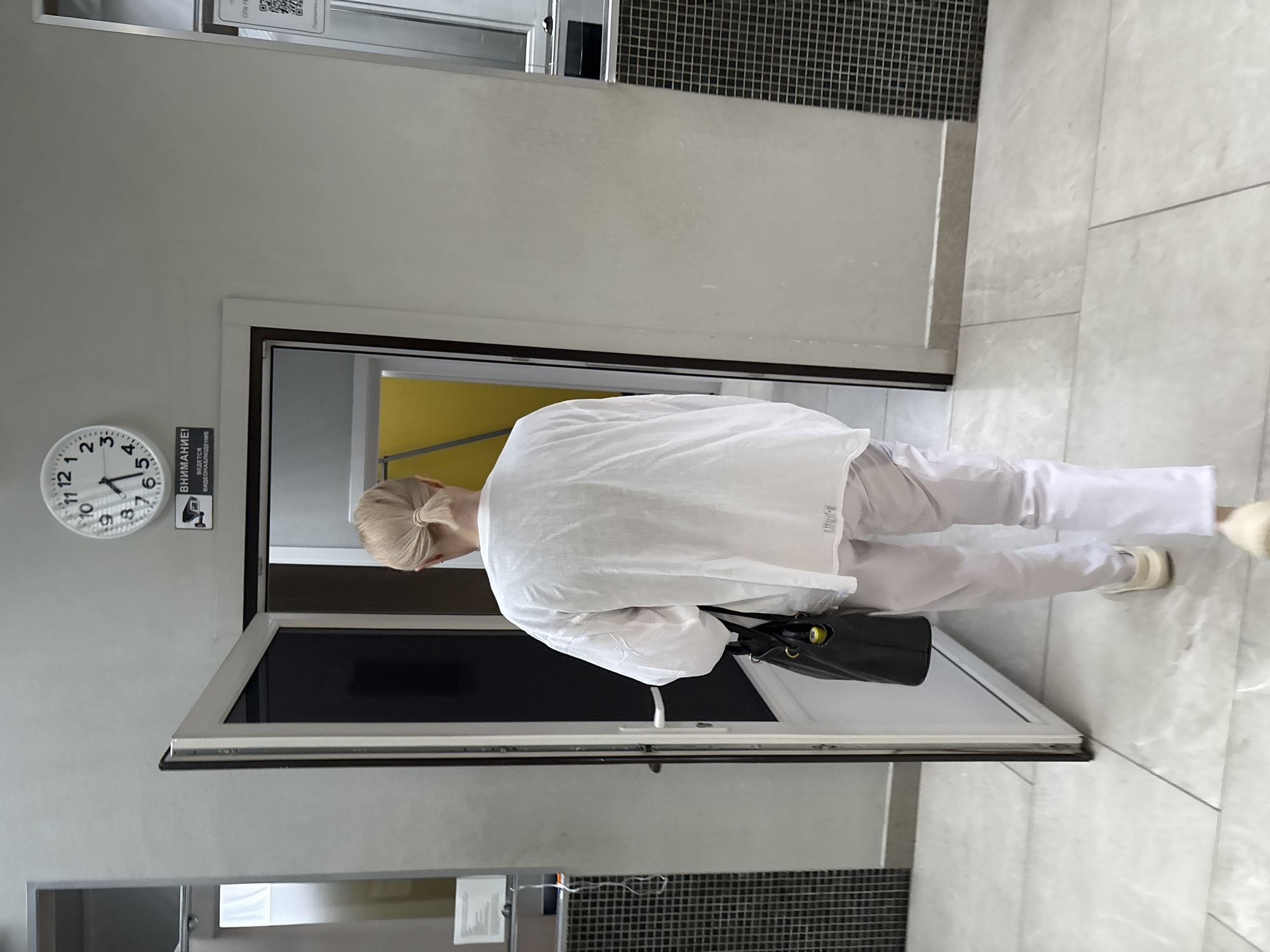 An adult woman with blonde hair pulled into a ponytail, wearing a white long-sleeved shirt and white pants, is walking away from the camera towards an open doorway. She is carrying a black bag over her left shoulder. She is wearing light-colored shoes. The doorway leads into a room with a yellow wall visible in the background. Above the doorway, a round clock with black numbers and hands is mounted on a grey wall. Below the clock, a black and white sign with an image of a security camera and Russian text, indicating video surveillance, is visible. The floor is tiled with a light-colored pattern. The scene appears to be indoors, possibly in a public building or institution. The location in the city of Saint Petersburg, Russia, is not specified by any visible context in the image.