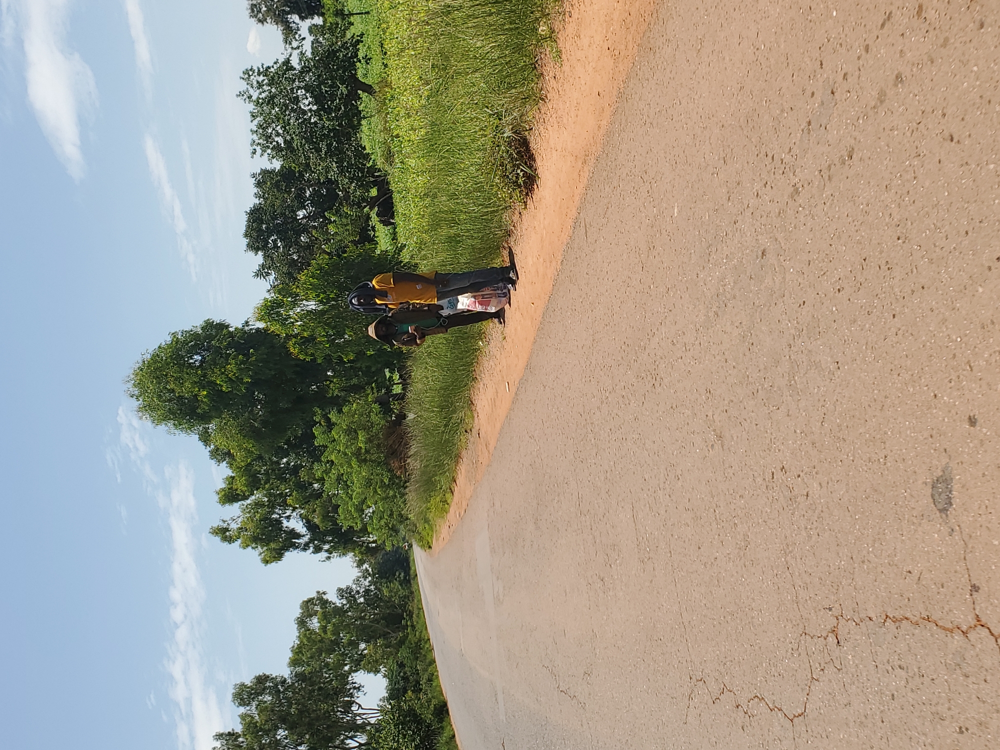 Two individuals stand on the side of a cracked asphalt road in a rural area of Mangu, Nigeria. The person on the left, wearing a brown hooded jacket and dark pants, holds a white and red plastic bag. The person on the right is dressed in a yellow t-shirt and blue jeans. Both are facing towards the right of the frame. The road curves to the left in the distance, bordered by dry, sandy earth and tall green grass. A dense line of trees and foliage forms the background. The sky above is a clear blue with scattered white clouds.