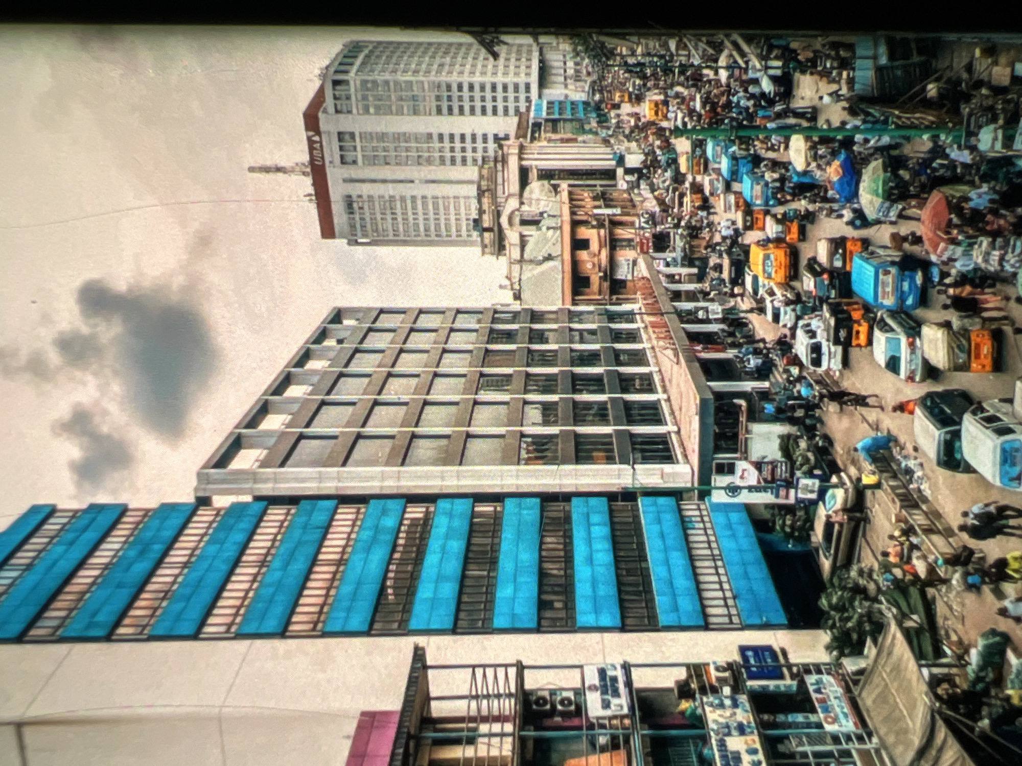 This is an aerial view of a bustling street scene in Lagos, Nigeria. The image depicts a dense urban environment with tall buildings lining both sides of a wide street packed with vehicles and pedestrians. In the foreground, to the left, is a tall, light-colored building with striking horizontal bands of bright blue windows. Adjacent to it is another, darker building with a grid-like pattern of windows.
Further down the street, a very tall, white skyscraper with the "UBA" logo is visible in the background, dominating the skyline. The street itself is filled with a variety of vehicles, including cars, buses, minibuses, and what appear to be auto-rickshaws or Keke Napeps. Many people are walking on the sidewalks and crossing the street, suggesting a busy commercial area.
On the lower levels of the buildings, storefronts and market stalls are visible, some with awnings providing shade. The overall impression is one of a vibrant and energetic city, with a high density of activity. The sky is overcast with scattered clouds. The lighting suggests it is daytime, though the sky is not clear blue.