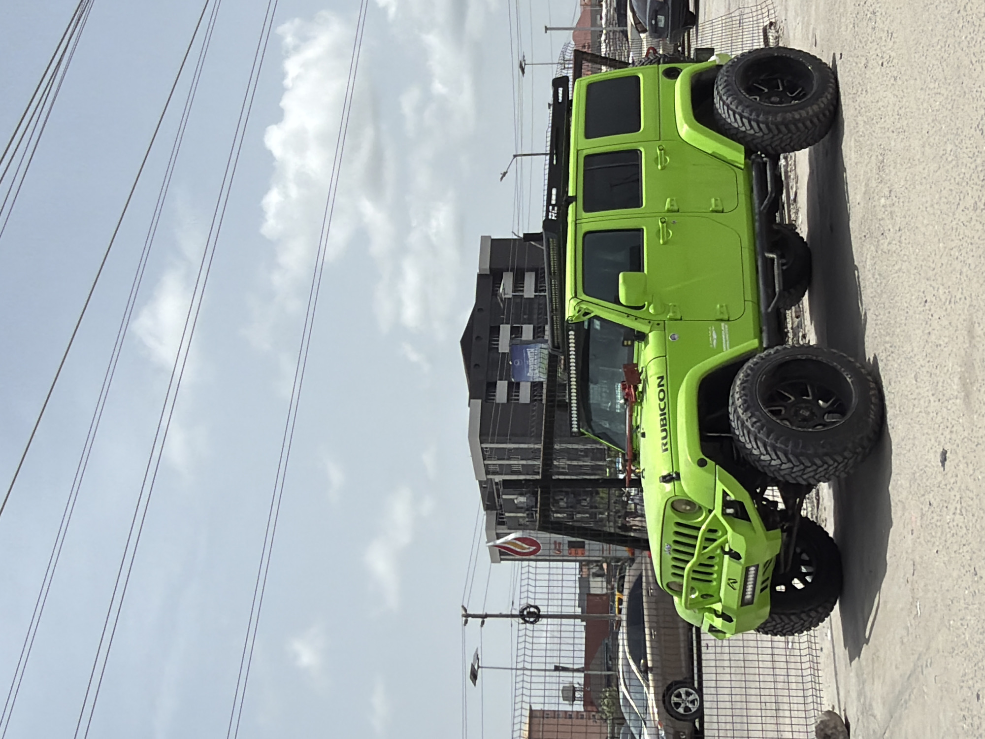 The image depicts a bright green Jeep Rubicon parked on a paved surface in what appears to be Igboefon, Nigeria. The car is heavily modified with large, black tires and a custom front bumper. Background details include buildings, power lines, streetlights, and other vehicles. A sign with a red and white logo is visible near the Jeep. The sky is partly cloudy. There are no people in the immediate view of the camera.