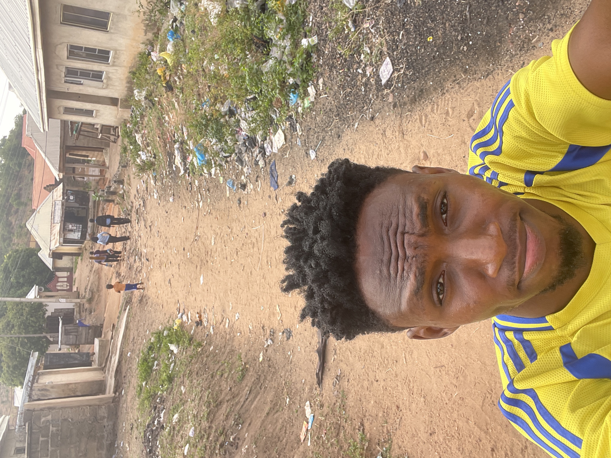 The original image is rotated 90 degrees clockwise. When viewed upright, it presents an outdoor scene in what appears to be a semi-urban or residential area, consistent with the provided context of Lokoja, Nigeria.
In the foreground, a Black man is taking a selfie, his face and upper torso occupying the right side of the frame. He has dark skin, short, dark curly hair, and a neatly trimmed beard. He is wearing a vibrant yellow short-sleeved athletic jersey, featuring distinctive blue stripes on the shoulders and sleeves that resemble the Adidas brand design. His expression is neutral and direct, with a slight furrow in his brow, and he is looking straight into the camera.
The background unfolds across a wide expanse of sandy, unpaved ground, which is noticeably strewn with various pieces of litter and debris, including plastic bags and wrappers. Interspersed with the dirt are patches of sparse green vegetation and weeds, also mixed with trash. To the left, several multi-story buildings are visible, some appearing to be in various stages of construction or showing signs of wear.
In the mid-ground, several other individuals can be seen. A small group of adults is standing together near one of the buildings, and further down the unpaved path, a lone child-like figure in an orange top and light-colored bottoms is walking. The scene is brightly lit by natural daylight, indicating a clear day. There is no clearly discernible text or branding visible on any objects or signs within the image, beyond the implied sportswear brand on the main subject's shirt.