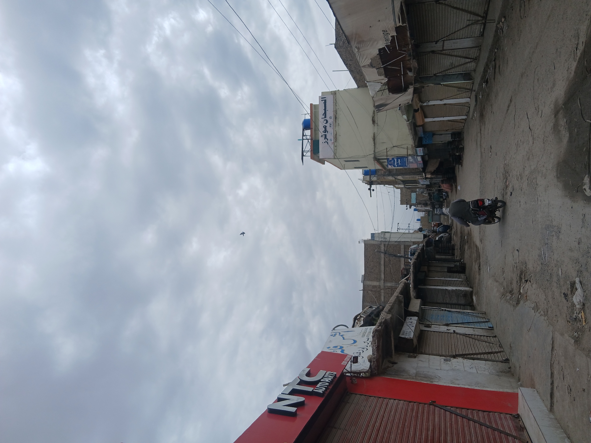 The image captures a street scene in an urban or semi-urban area, likely in Quetta, Pakistan, under an overcast sky. The sky is largely covered with grey and white clouds, indicating a cloudy day, with a small dark bird visible in the upper part of the frame.
The street itself appears to be unpaved or poorly maintained, characterized by dust and uneven surfaces. Buildings line both sides of the street, typically two to three stories high, with several shopfronts. Many of these shops have their metal roller shutters pulled down, suggesting they are closed or not actively trading.
In the foreground, on the left side of the street, a prominent red and white sign for "NTC AUTO PARTS" is clearly visible. Further down the street, on a lighter-colored building, another sign displays text in Urdu or Arabic, including "الحصون" (Al-Husun) and "موبائل" (Mobile), along with phone numbers (e.g., 030 000xxxx, 030 000xxxx). Additional signs with similar script are partially visible on other buildings.
Numerous utility poles and overhead power lines crisscross the view, indicative of a developed area. On the rooftops of some buildings, blue water tanks can be seen. A single person on a motorcycle is captured in the middle-ground, riding down the street, facing away from the viewer. The overall atmosphere suggests a commercial district, possibly a market area, during an ordinary, overcast day.