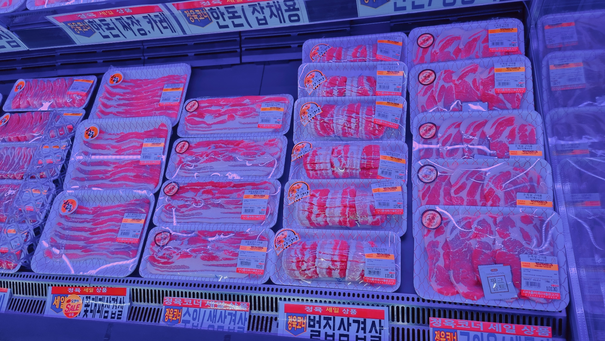The image shows a refrigerated display case in a grocery store, filled with packages of raw meat. The meat is thinly sliced and arranged on white polystyrene trays, wrapped in clear plastic. Each package has a price tag and label in Korean. The shelves are lined with metal grates, and signs in Korean advertising the meat are visible below. The packages are predominantly red, indicating different cuts of pork. The packages display the brands of "더다음마트(더좋은고기)" and "Premium". The shelves are likely located in a grocery store in Dunnae-myeon, South Korea.