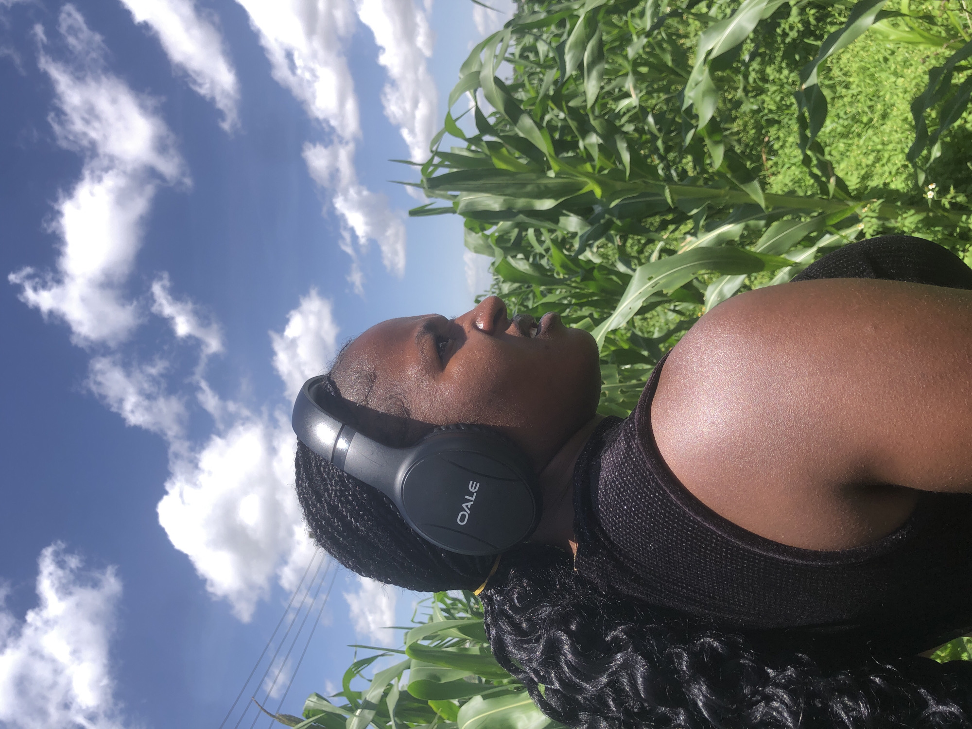 A medium shot captures a young Black woman from the shoulders up, in profile, facing right. She has dark braided hair that cascades down her back and is wearing black over-ear headphones with the brand name "OALE" in white lettering on the right earcup. She is wearing a black sleeveless top. Her skin is dark and appears slightly dewy, suggesting sweat or humidity.
The background is a lush, green cornfield under a bright blue sky dotted with white, fluffy clouds. The corn stalks are tall and vibrant, indicating a healthy crop. In the far distance, to the left of the frame, power lines are visible against the sky, suggesting proximity to a more developed area. The overall setting is rural, possibly agricultural land. Given the context provided about Gwafan, Nigeria, this scene depicts the woman in what appears to be an agricultural area within or near the city.