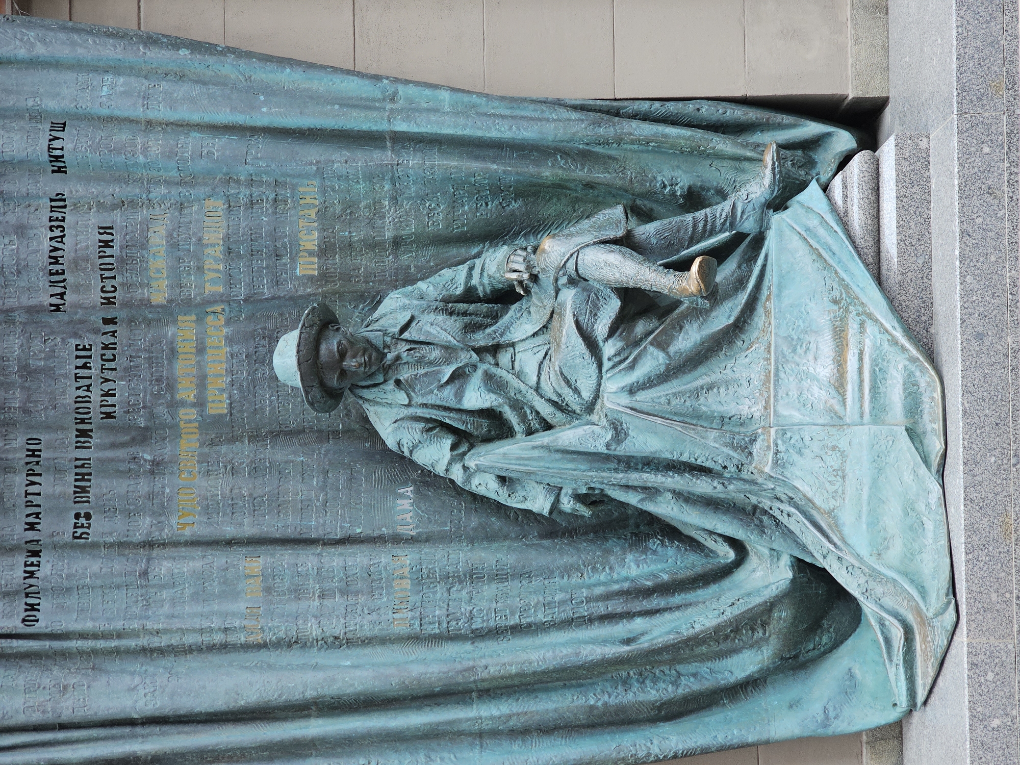 Here's a concise description of the image:
The image depicts a bronze relief sculpture mounted on a stone wall. The sculpture features the figure of a man in a hat, lying down, seemingly covered by a large draped cloth. The cloth appears to be part of the sculpture itself and is inscribed with Cyrillic text in various sizes and styles. The text is partially highlighted in gold color. Based on the text content and style, it is likely a memorial or historical marker located in Moscow, Russia.