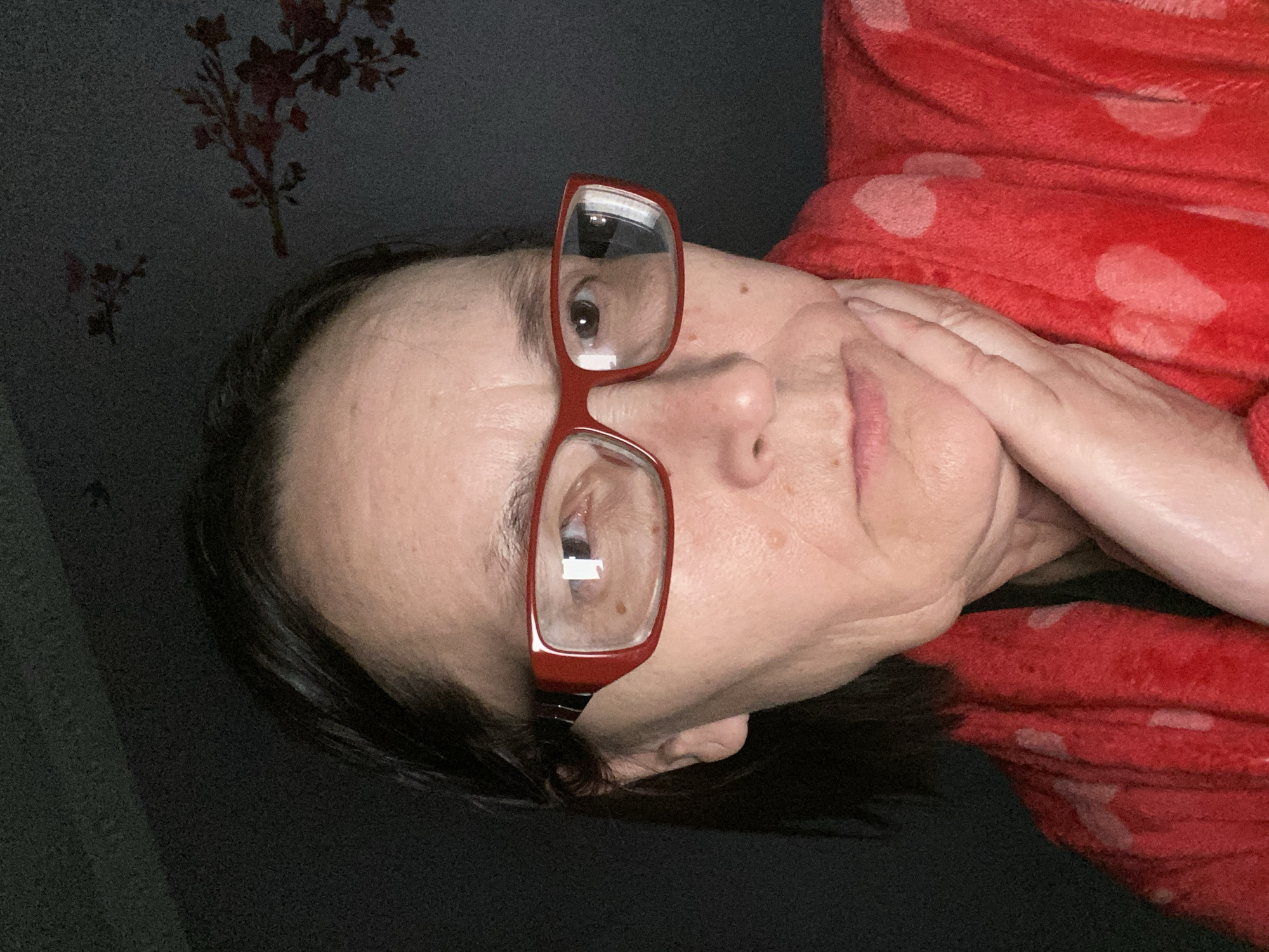 A woman with dark hair and red-framed glasses is positioned at an angle in the frame. Her hand is touching her face near her chin. She is wearing a red robe with white polka dots. The background consists of a dark gray wall with red floral patterns. The image does not provide enough information to determine the location in Novokuznetsk, Russia.