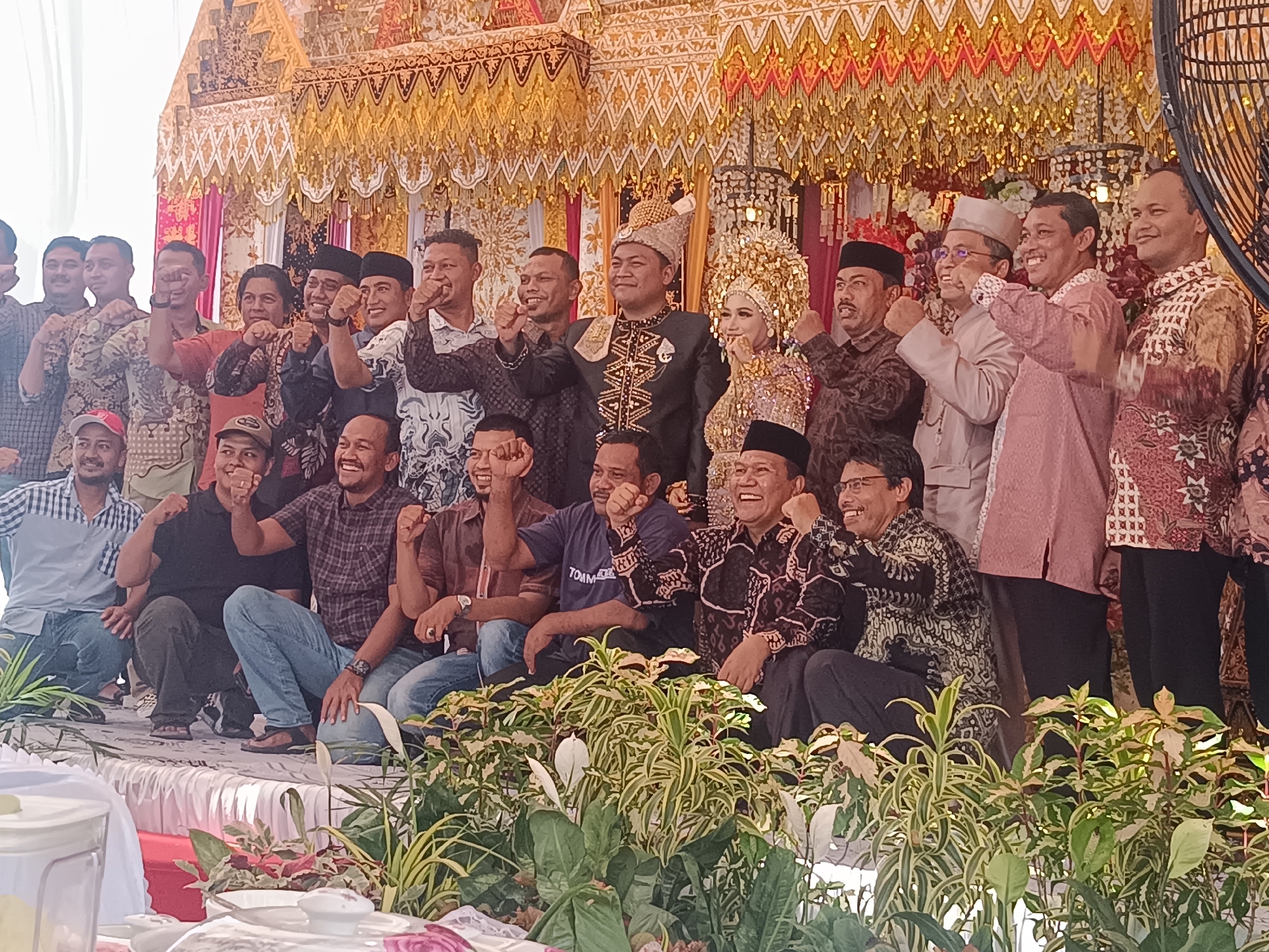 The image shows a group of people, including a bride and groom in traditional attire, posing for a photo at what appears to be a wedding celebration. They are located in Lhokseumawe, Indonesia. The group is standing and sitting on a raised platform that is adorned with flowers and foliage. Behind the group is an elaborate, golden-colored decorative backdrop, likely part of the wedding venue. Many of the people in the group are raising their fists in the air, possibly in a celebratory gesture. The bride is wearing an ornate headdress and the groom is dressed in a traditional suit with a decorative headdress. The scene suggests a festive and communal atmosphere.