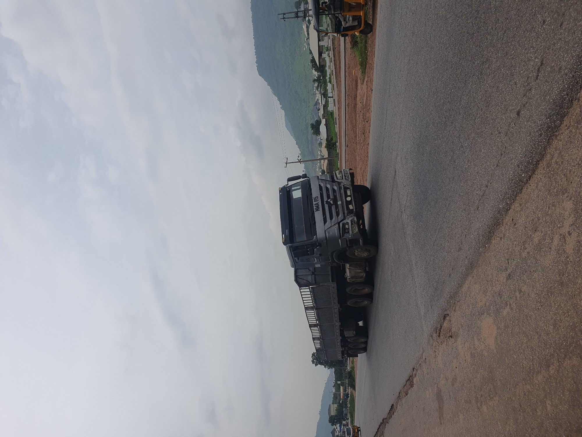 A large, grey flatbed truck, with a metal railing around the cargo bed, is driving on an asphalt road in Lokoja, Nigeria. The truck has "HWA 45" written on the front. To the left of the truck, a smaller yellow three-wheeled vehicle, known as an "okada" or "Keke NAPEP", is also visible. In the background, there are green hills with scattered buildings and trees, and the sky is overcast with clouds. The road is a mixture of well-maintained asphalt and rougher patches with exposed dirt.
